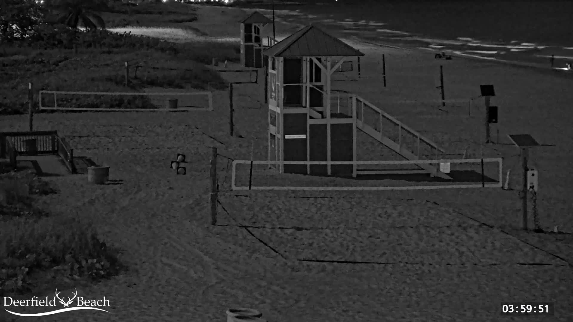 A dark, clear night reveals a sandy beach with two volleyball courts, two observation towers, and the ocean with visible waves along the right edge.