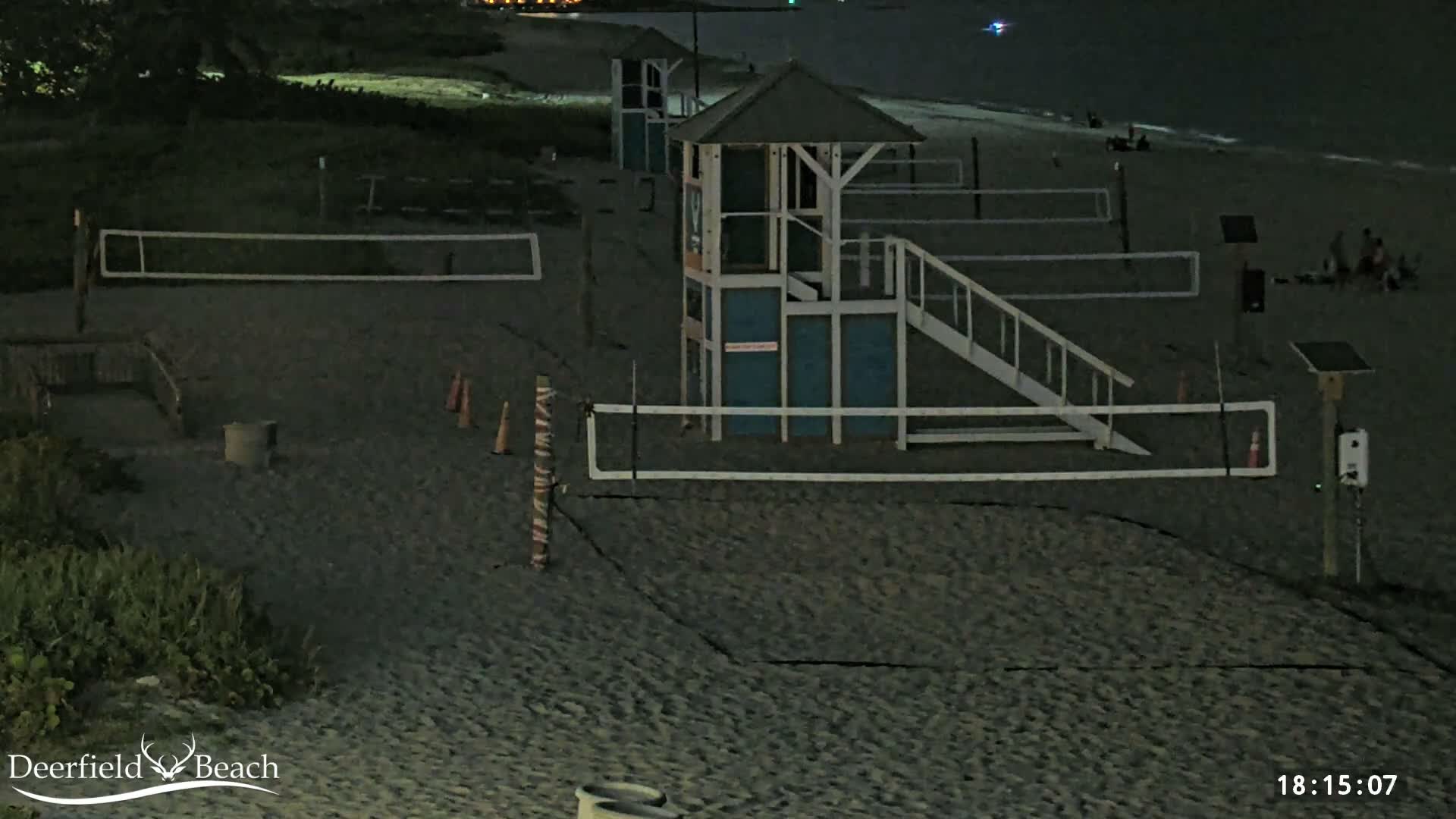 A dark, clear night reveals a sandy beach with two volleyball courts, two observation towers, and the ocean with visible waves along the right edge.