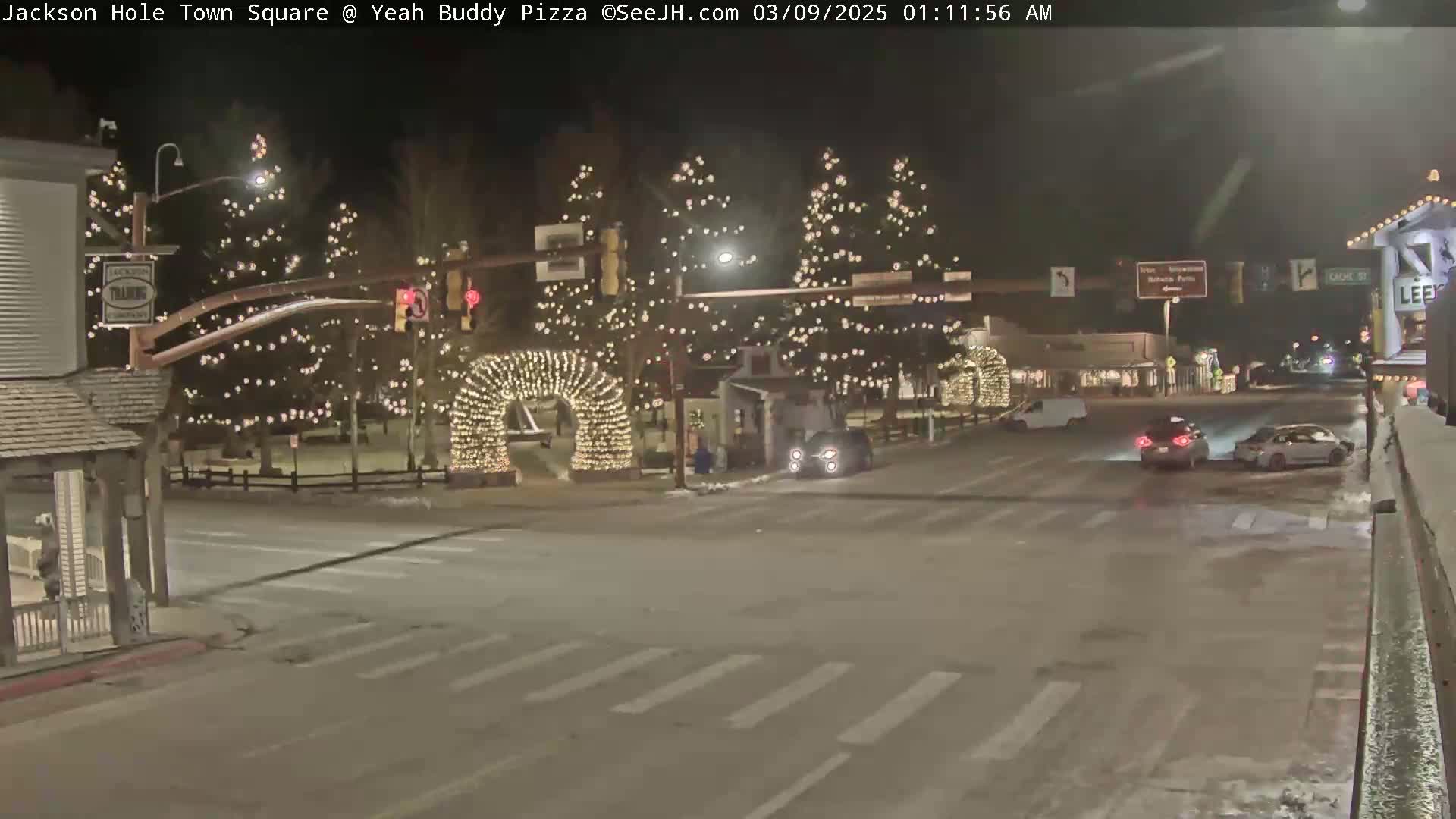 A snow-covered town square at night, illuminated by Christmas lights strung on trees and arches, with a few cars at an intersection.