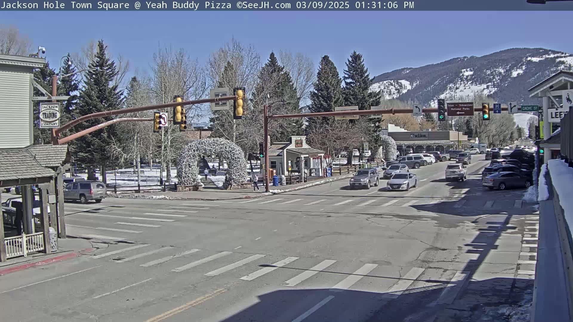 A sunny day in a town square shows a street intersection with cars, pedestrians, and buildings, with mountains in the background and some snow on the ground.