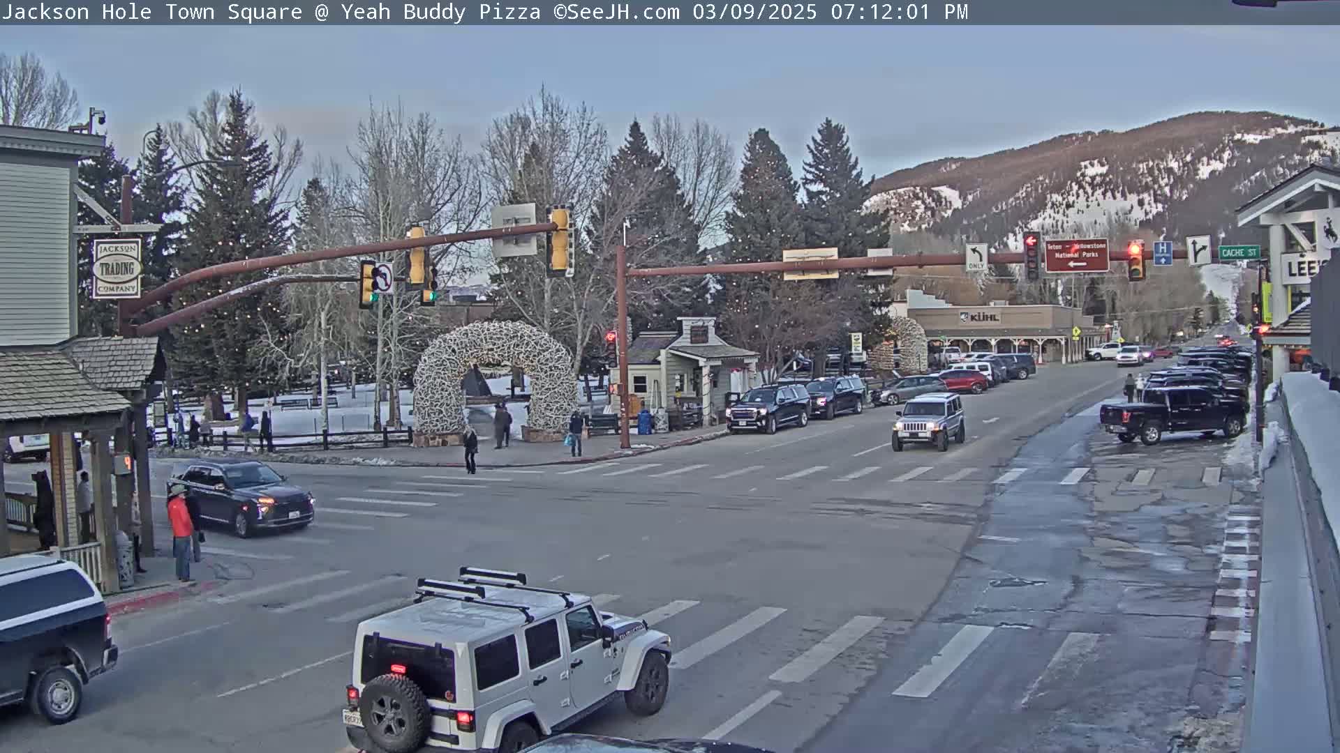 A lightly snow-covered town square with cars at an intersection, pedestrians on sidewalks, and a mountain range in the background.