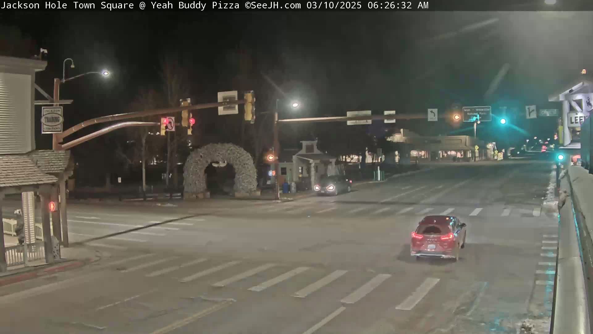 A nighttime view of a mostly empty town square intersection with traffic lights, a few cars, and some buildings, under clear skies.