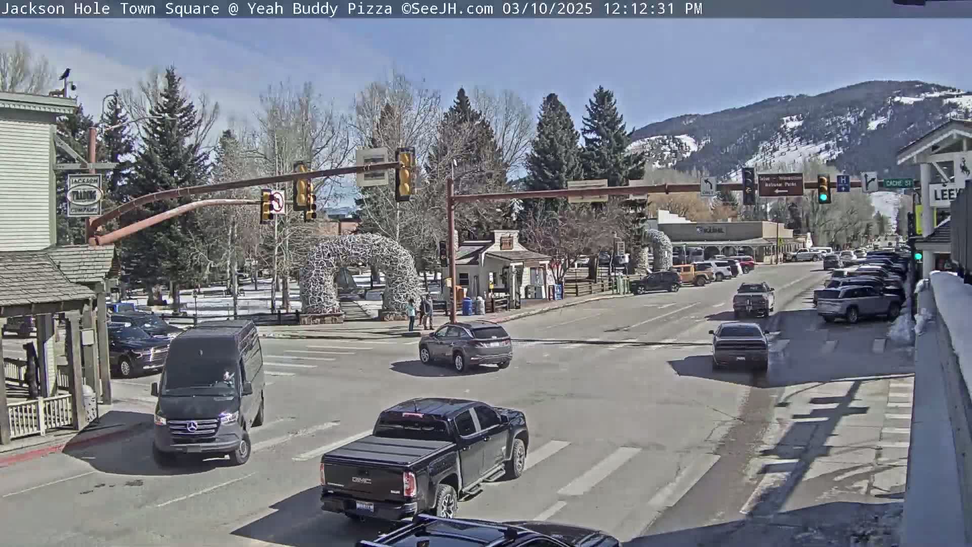 A sunny day in a town square shows several vehicles at a crossroad, with mountains in the background and pedestrians near a metal sculpture.