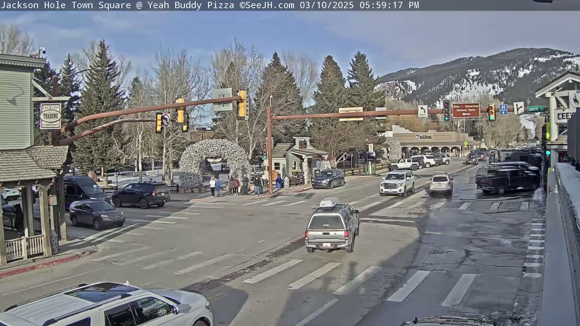 A sunny day in a town square shows a traffic intersection with several cars, pedestrians, and buildings, with snow-capped mountains in the background.