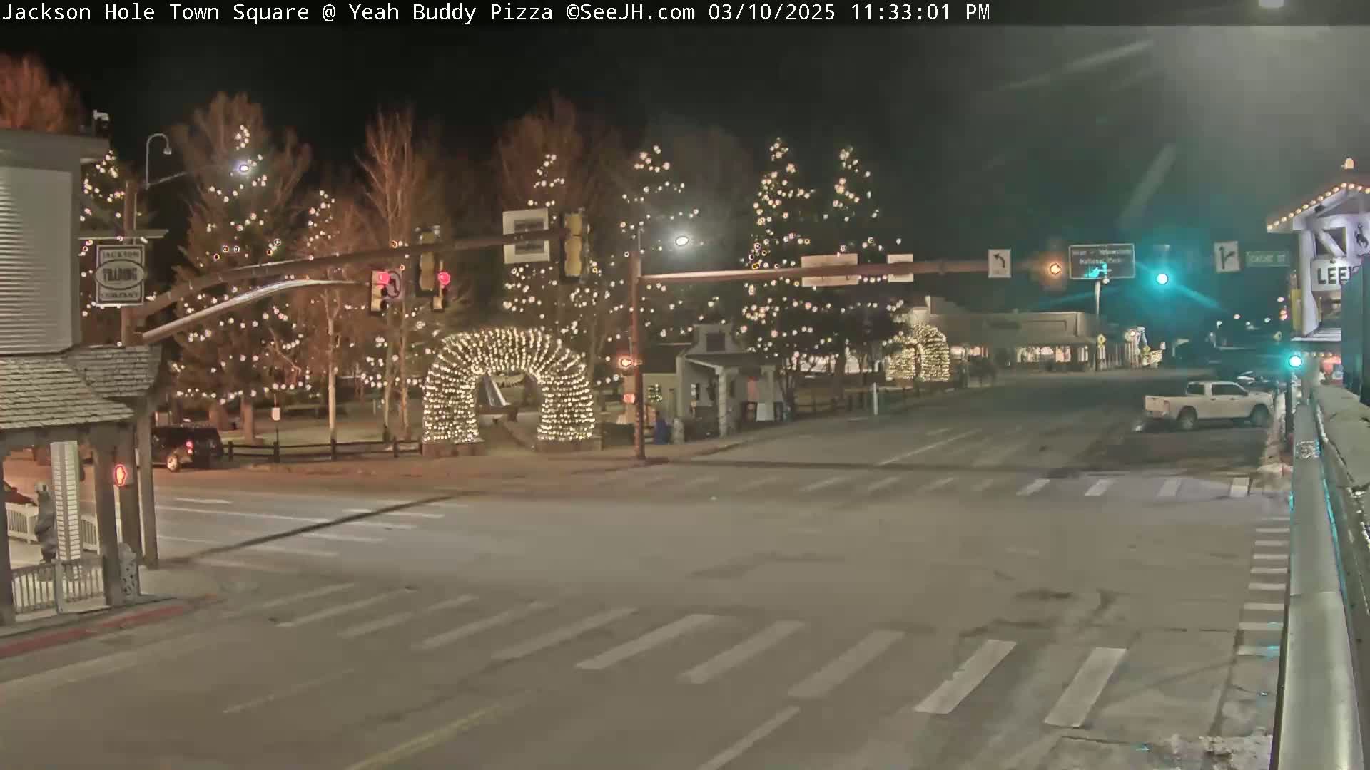 A nighttime view of a town square decorated with string lights on trees and arches, with a few vehicles on the road.