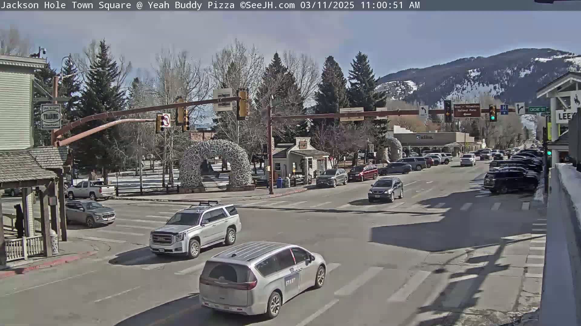 A sunny day in a town square with cars at a crosswalk, buildings lining the street, and mountains in the background.