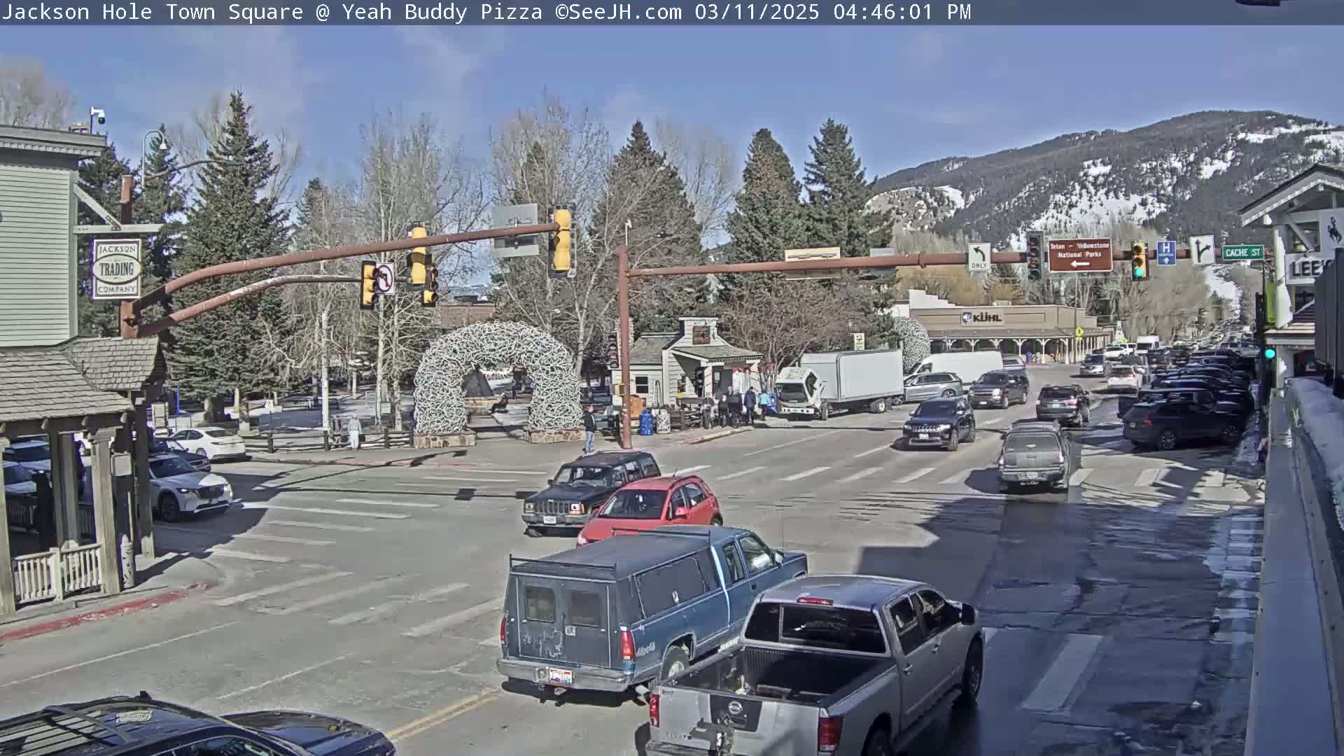 A sunny day in a town square shows several vehicles at a multi-lane intersection, with mountains visible in the background and trees lining the streets.
