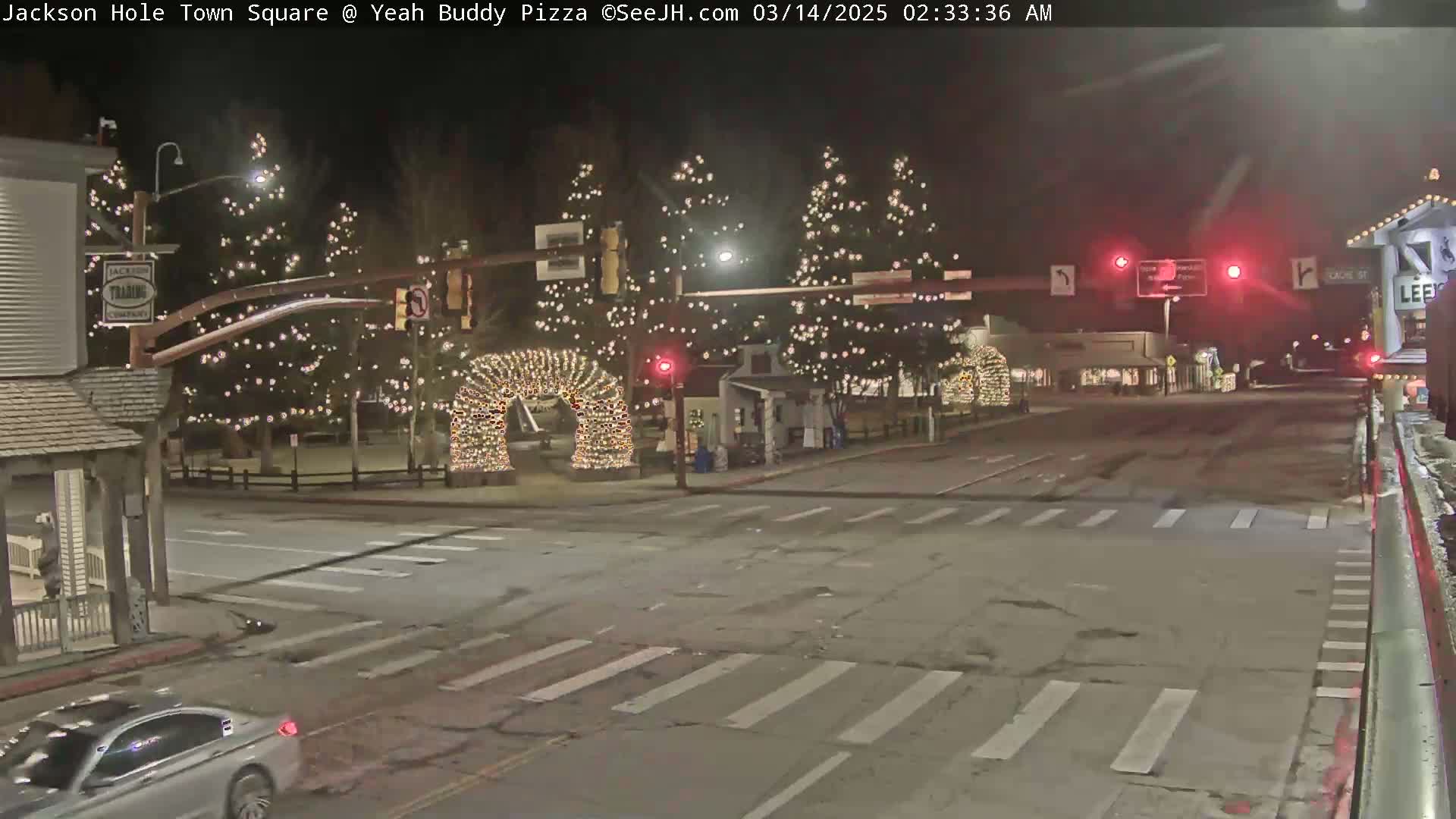 A nighttime street scene shows a town square decorated with Christmas lights, including illuminated arches, trees, and buildings.