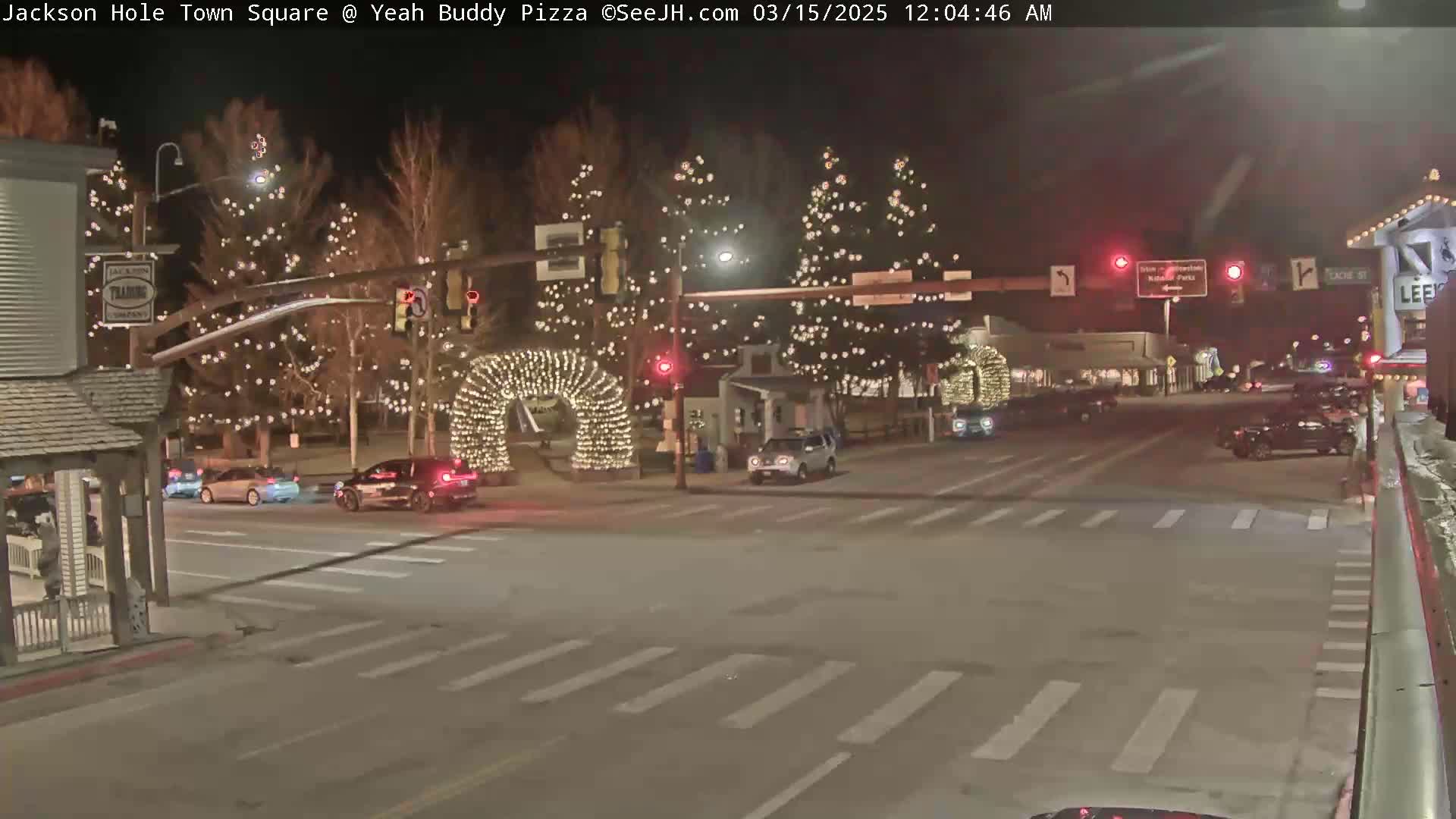 A nighttime scene of a town square with several cars at an intersection, illuminated by string lights on trees and archways.