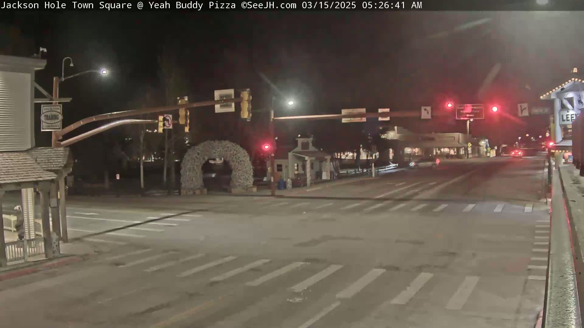 A nighttime view of a mostly empty town square intersection with traffic lights, a large antler arch, and a few buildings visible under a dark sky.