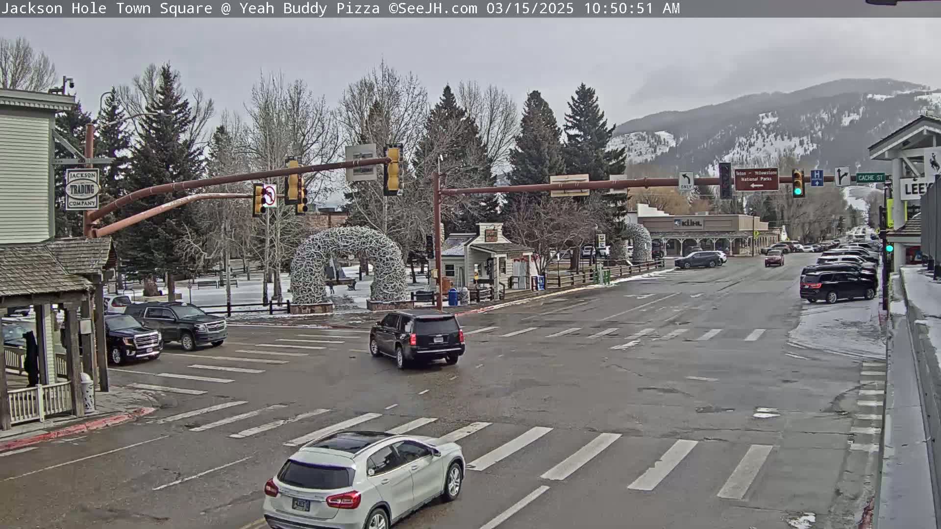 A town square with several cars at an intersection under an overcast sky, with snow visible on the ground and mountains in the background.