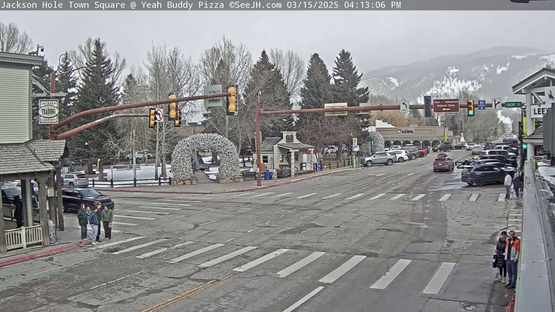 A town square with several cars parked along the street, a few pedestrians crossing a crosswalk, and a mostly overcast sky.