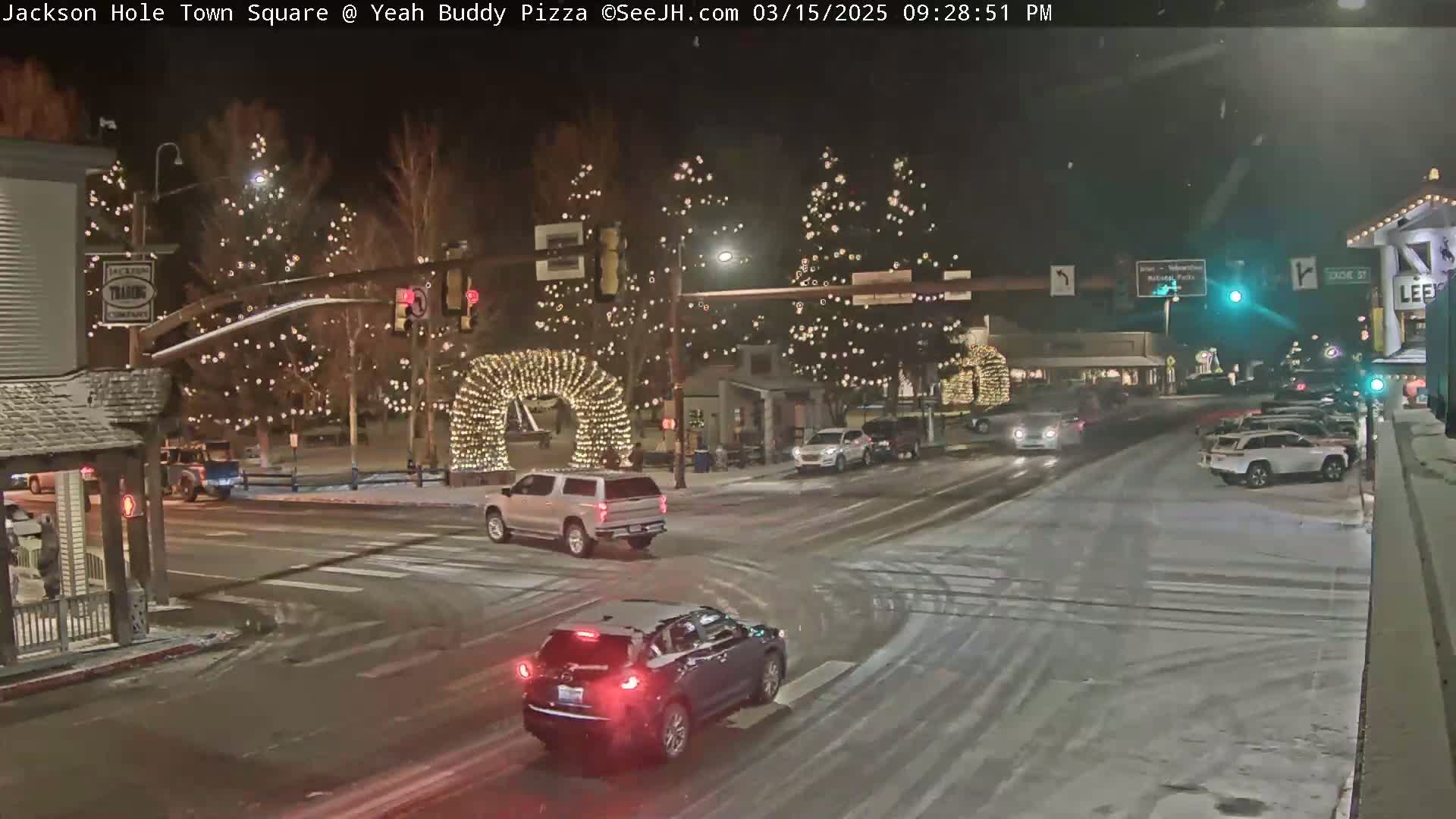 A snow-covered town square at night, illuminated by string lights on trees and an archway, with several vehicles driving through the intersection.