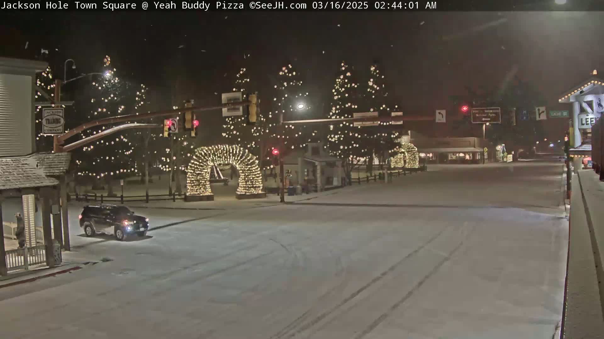 A snow-covered town square at night, illuminated by Christmas lights strung on trees and an archway, with a dark-colored SUV driving through the square.