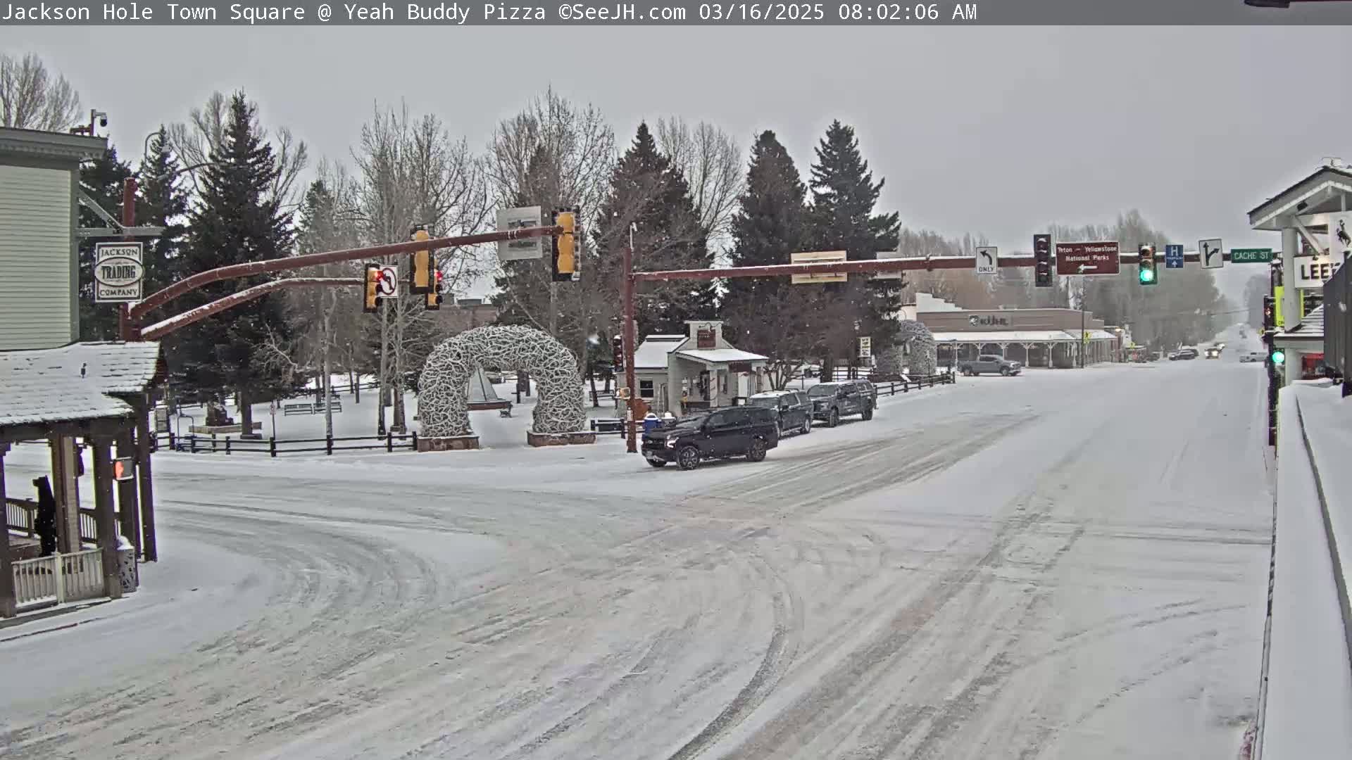 A snow-covered town square intersection with several cars and buildings, under light snowfall.