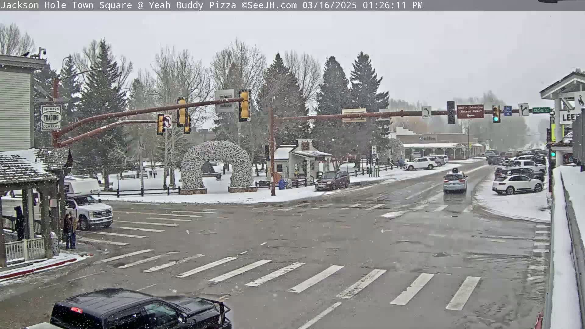 A snow-covered town square with cars at an intersection under a light snowfall.