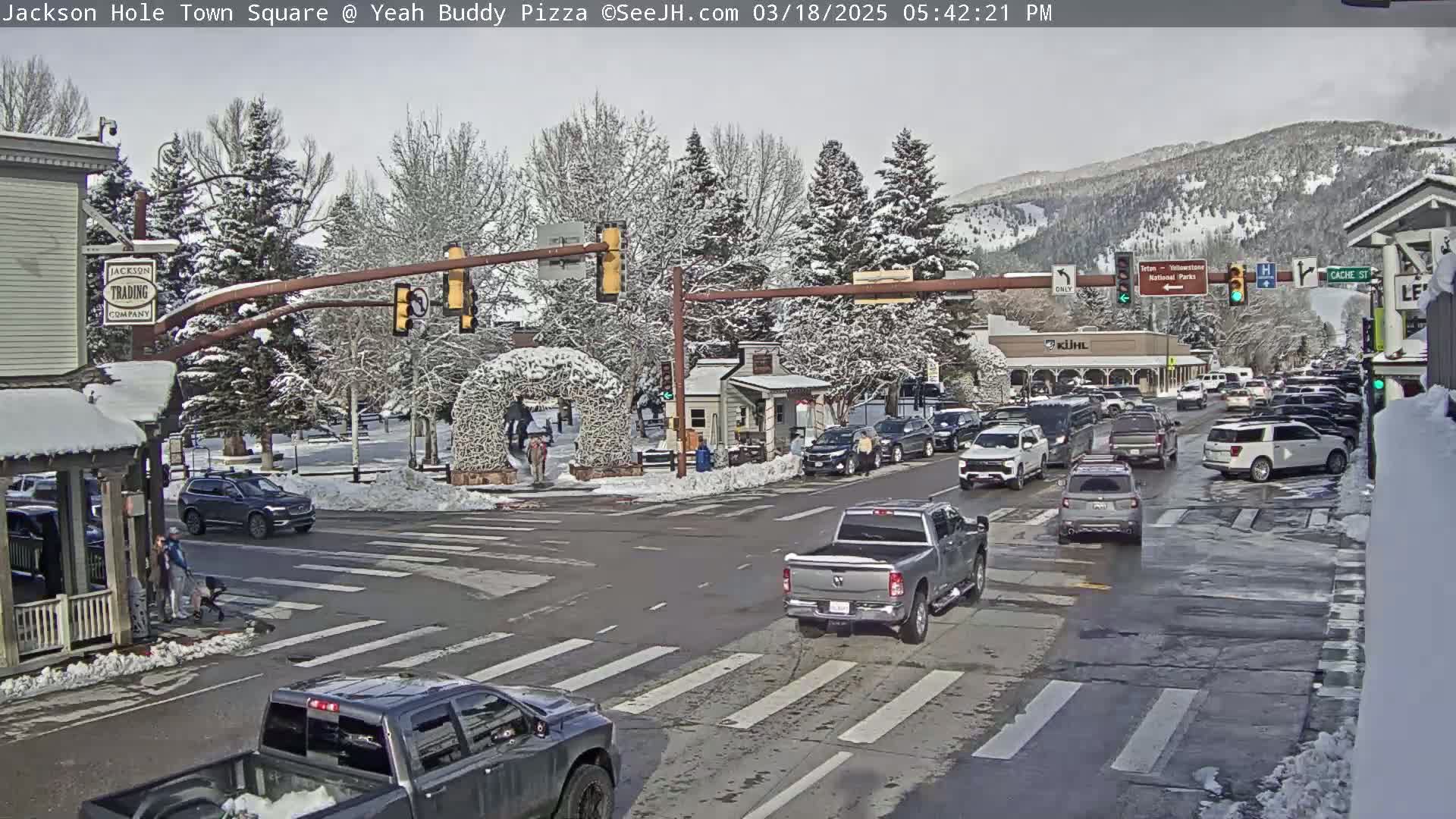 A snow-covered town square intersection shows several vehicles stopped at traffic signals, with pedestrians crossing the street and a background of snow-dusted trees and mountains.