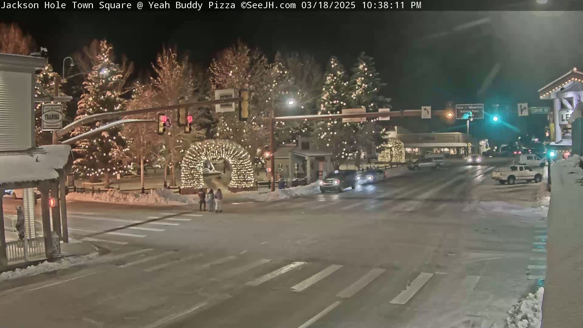 A snow-covered town square at night, illuminated by string lights on trees and an archway, with several vehicles at an intersection.