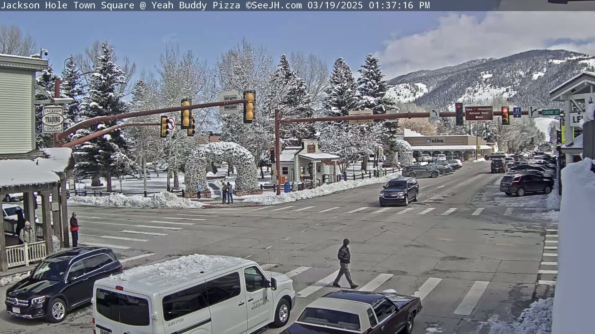 A snow-covered town square with cars parked along the street and pedestrians crossing the road under a mostly sunny sky, with mountains in the background.