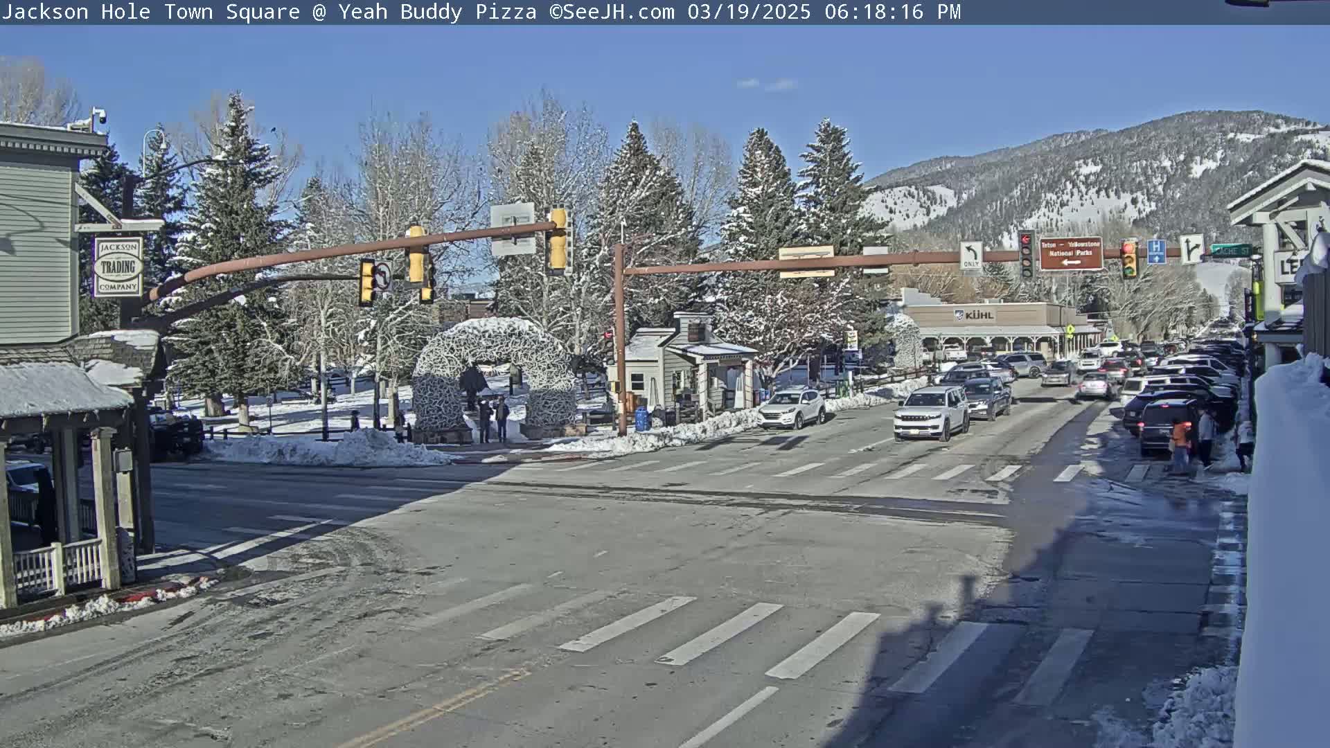 A sunny day in a snow-dusted town square, showing a street intersection with cars and pedestrians, and snow-covered trees and buildings in the background.