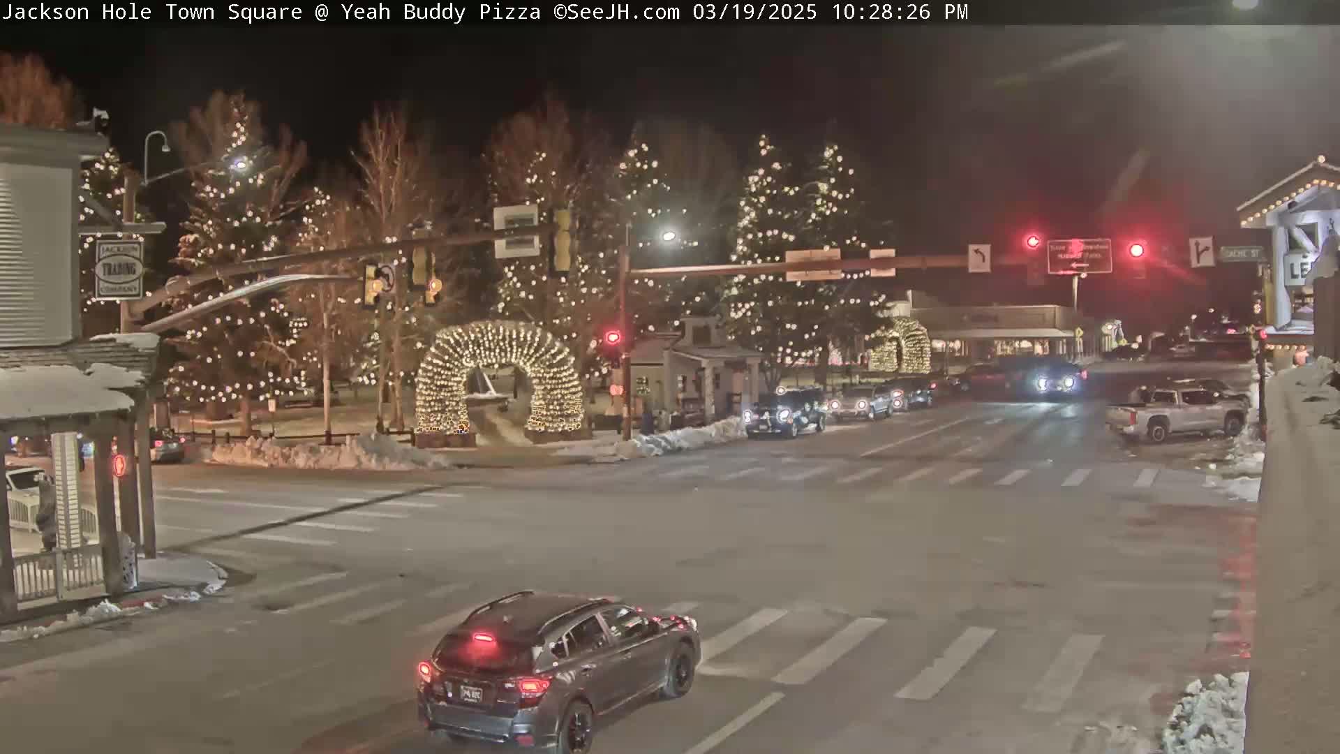 A nighttime street scene in a town square, decorated with Christmas lights on trees and an archway, shows cars at an intersection with some snow on the ground.