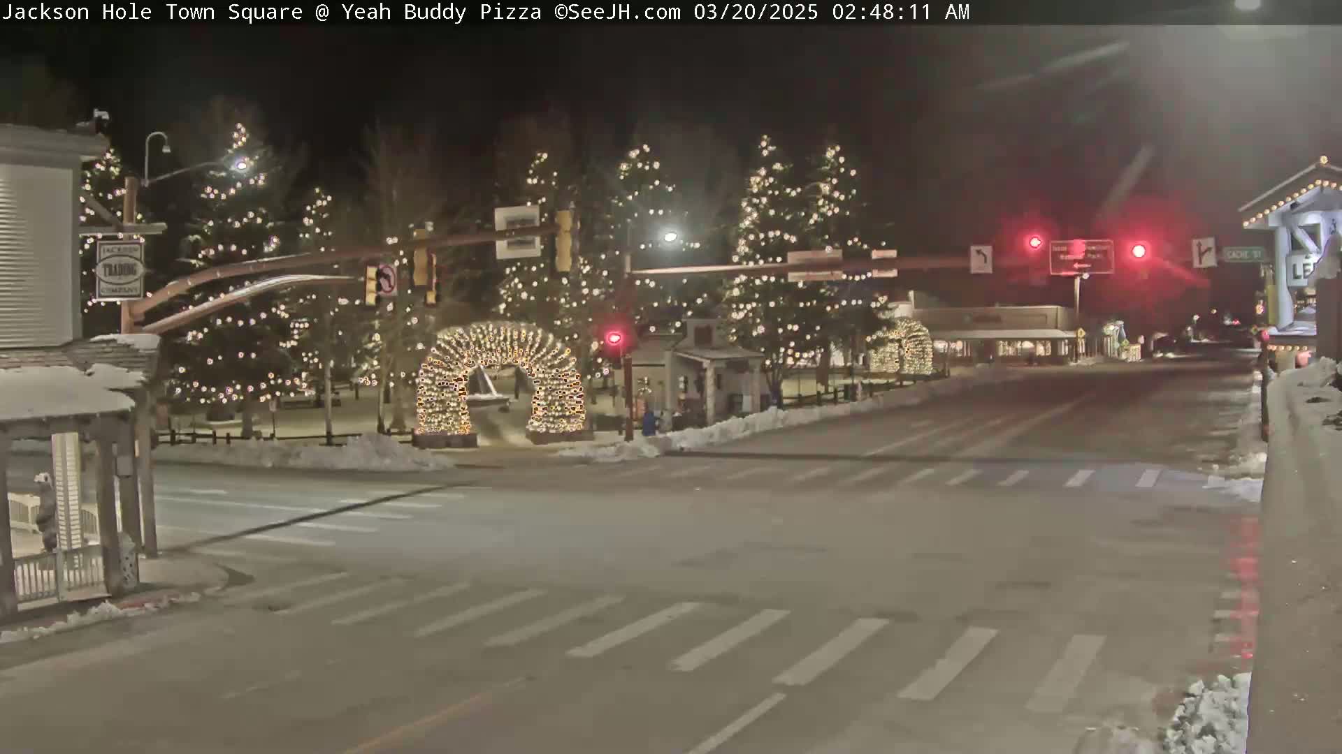 A snow-covered town square at night, illuminated by Christmas lights strung on trees and an archway, with traffic lights visible in the background.
