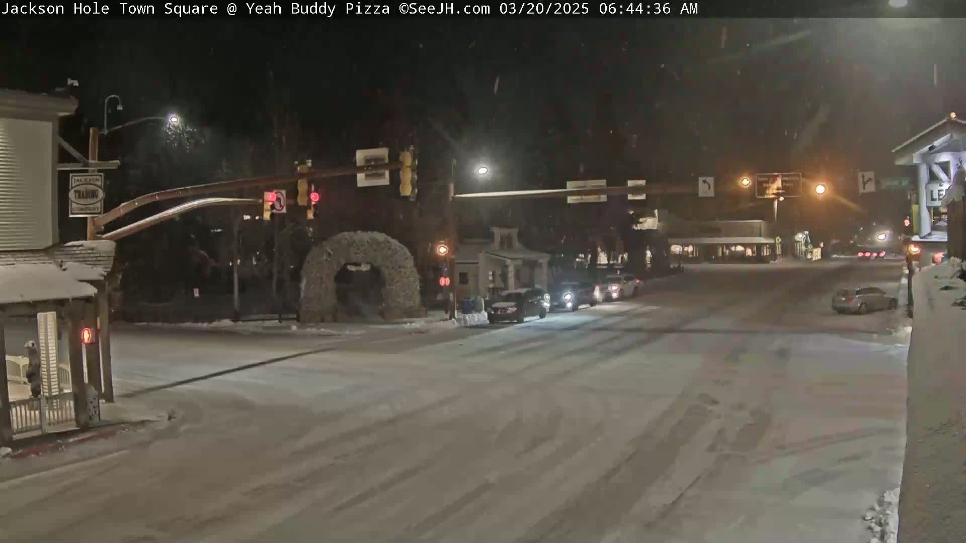 A snow-covered town square at night, with a few cars driving through a lightly trafficked intersection under illuminated streetlights.