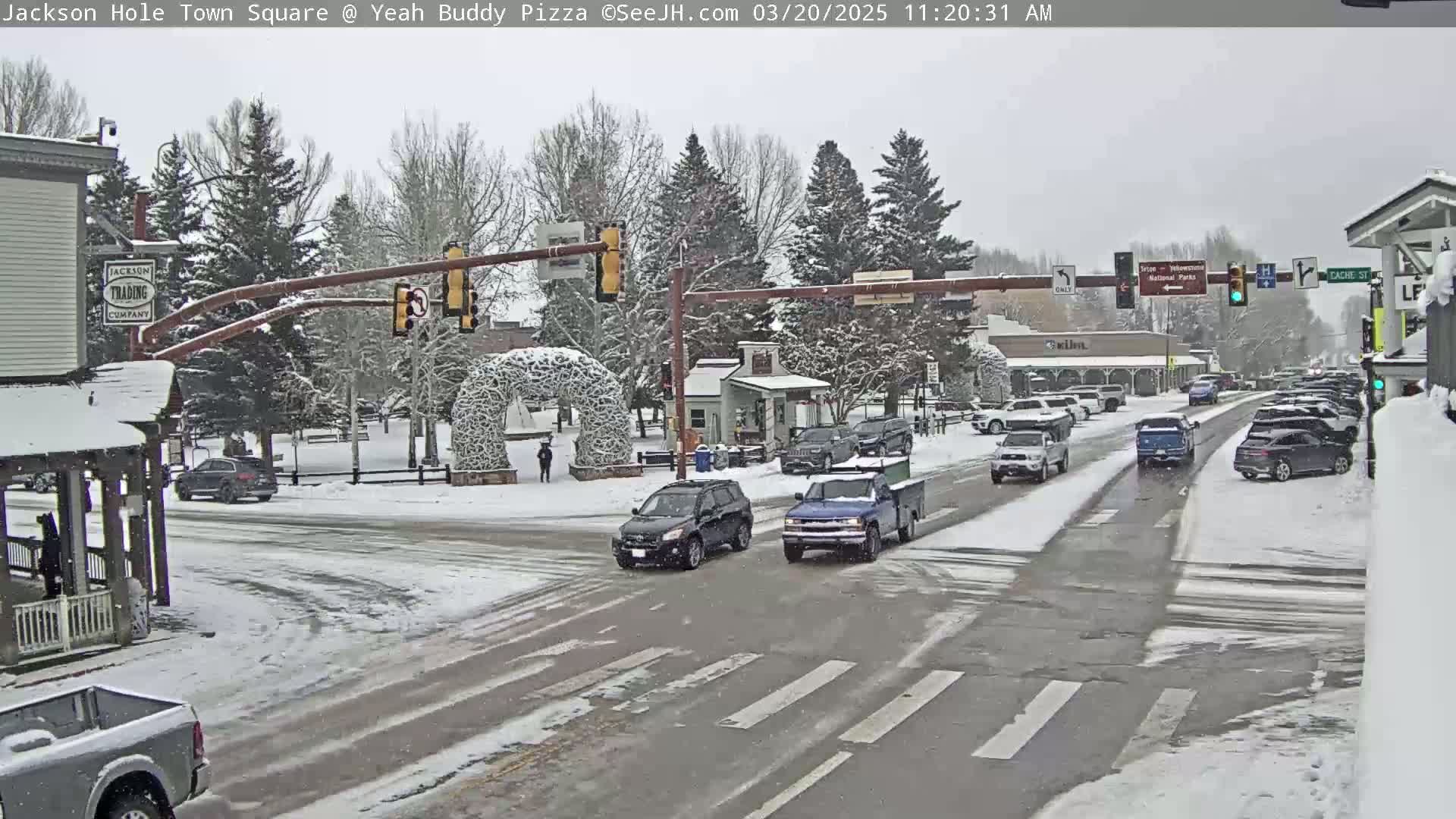 A snow-covered town square with cars driving on a street and a pedestrian walking.