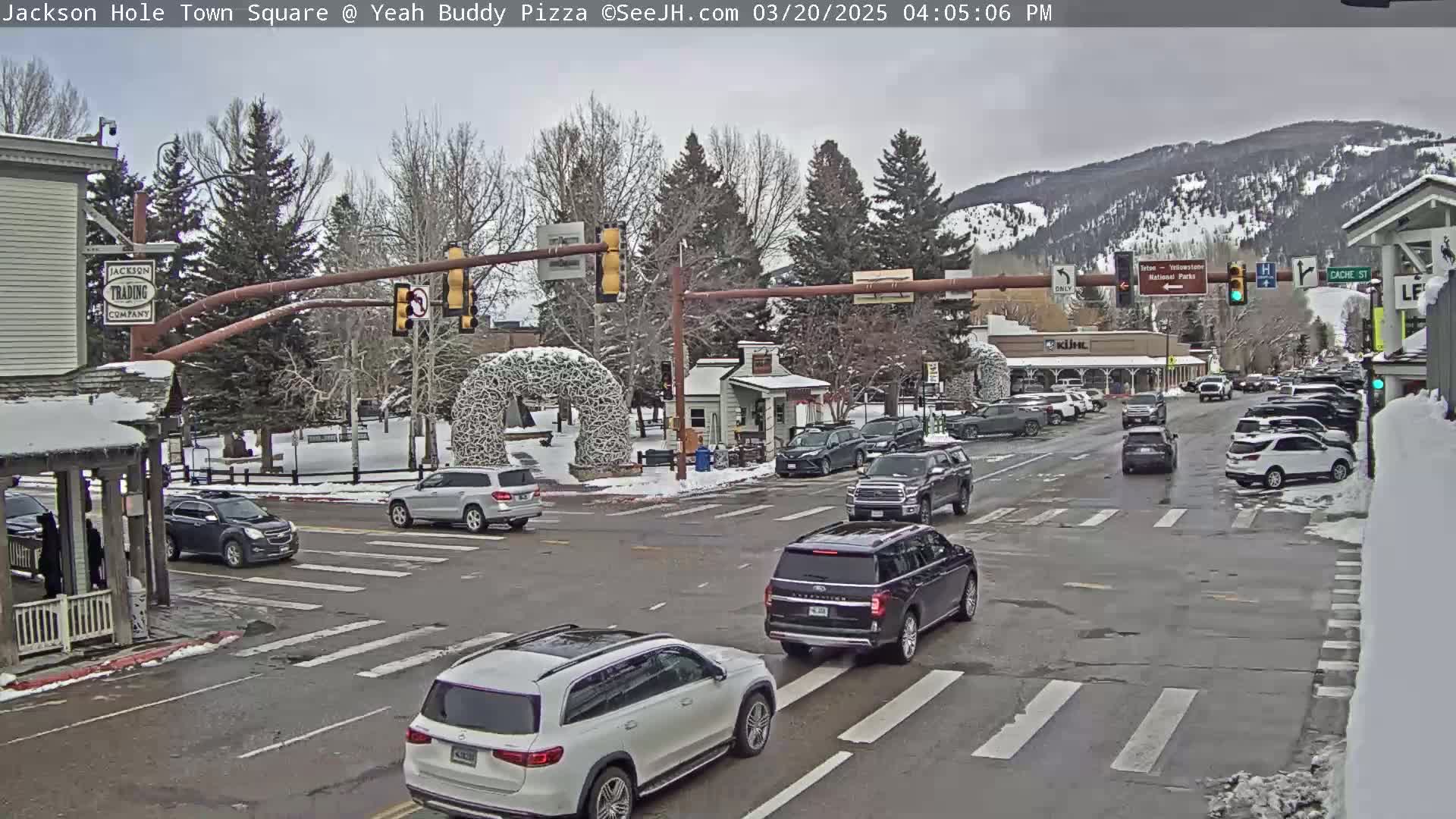 A town square intersection with several cars, snow on the ground, and a mountain range in the background under a cloudy sky.