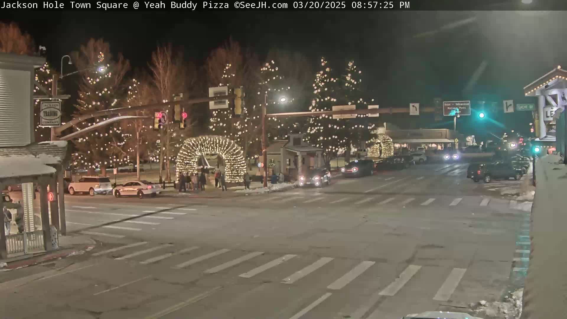 A nighttime scene of a town square with Christmas lights adorning trees and an archway, cars driving on the road, and some people standing in the square.