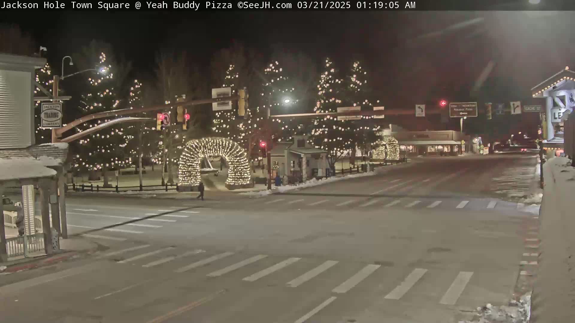 A nighttime scene of a town square with Christmas lights illuminating trees and an archway, with some snow on the ground.