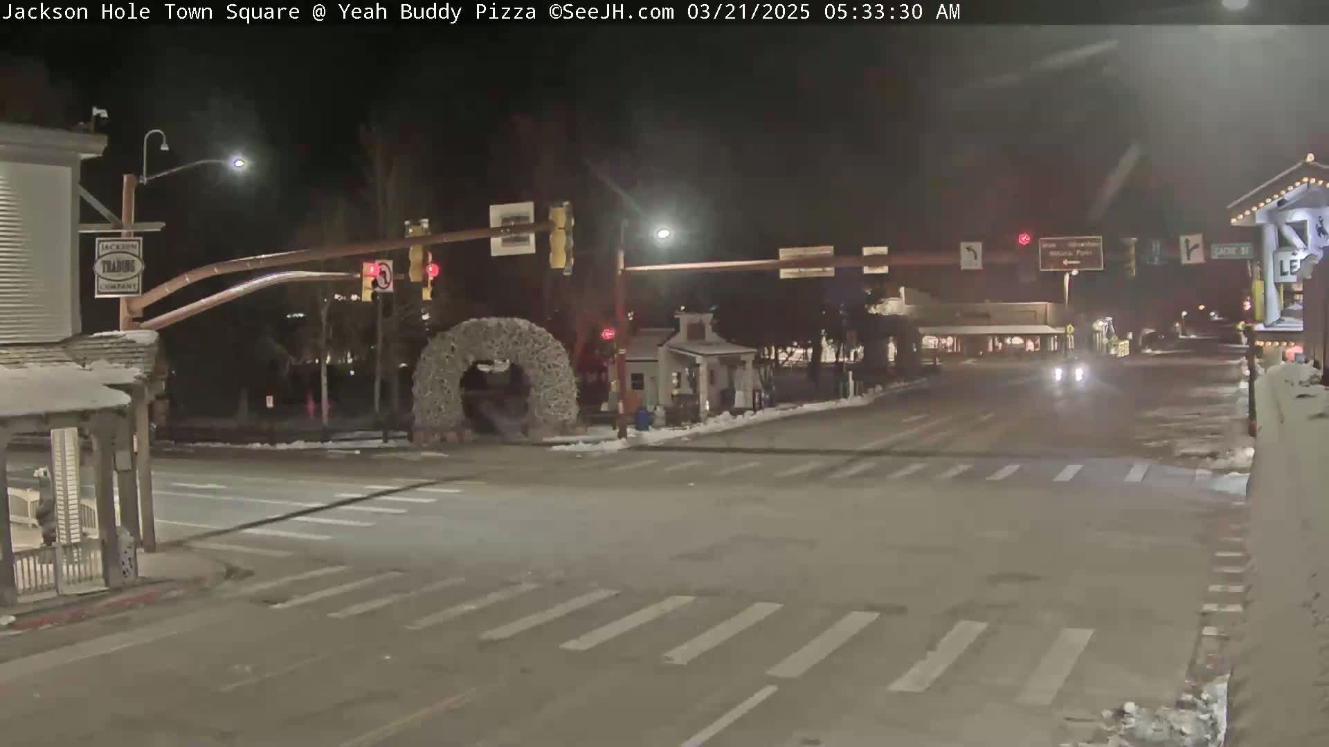 A nighttime view of a snow-covered town square intersection with traffic lights, a few cars, and a large decorative wreath.