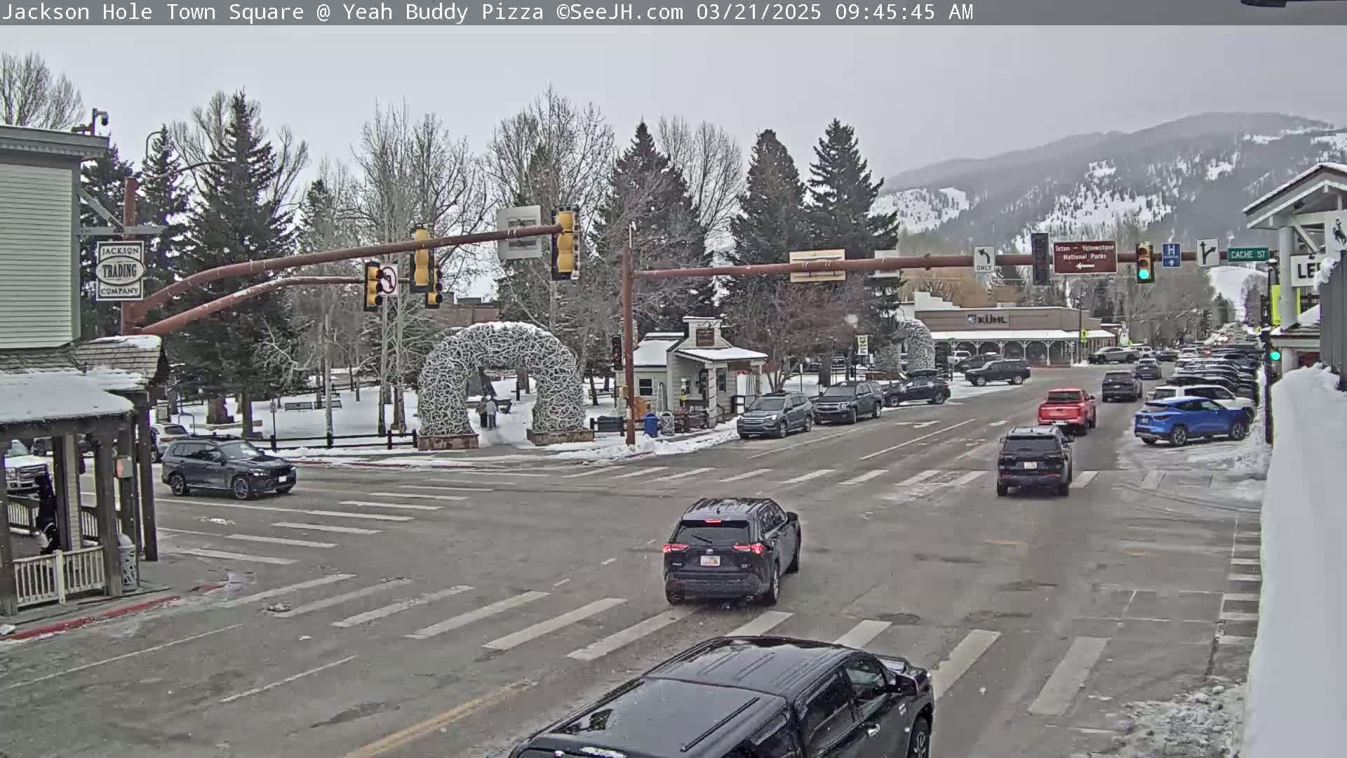 A snow-dusted town square shows several vehicles at an intersection under an overcast sky, with mountains visible in the background.