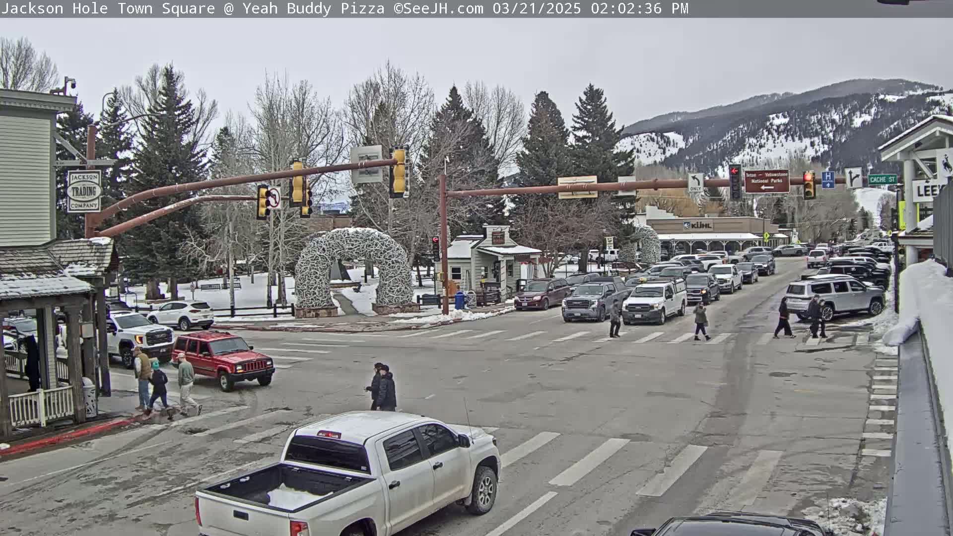 A town square intersection with several cars and pedestrians, under a cloudy sky, with snow on the ground and mountains in the background.