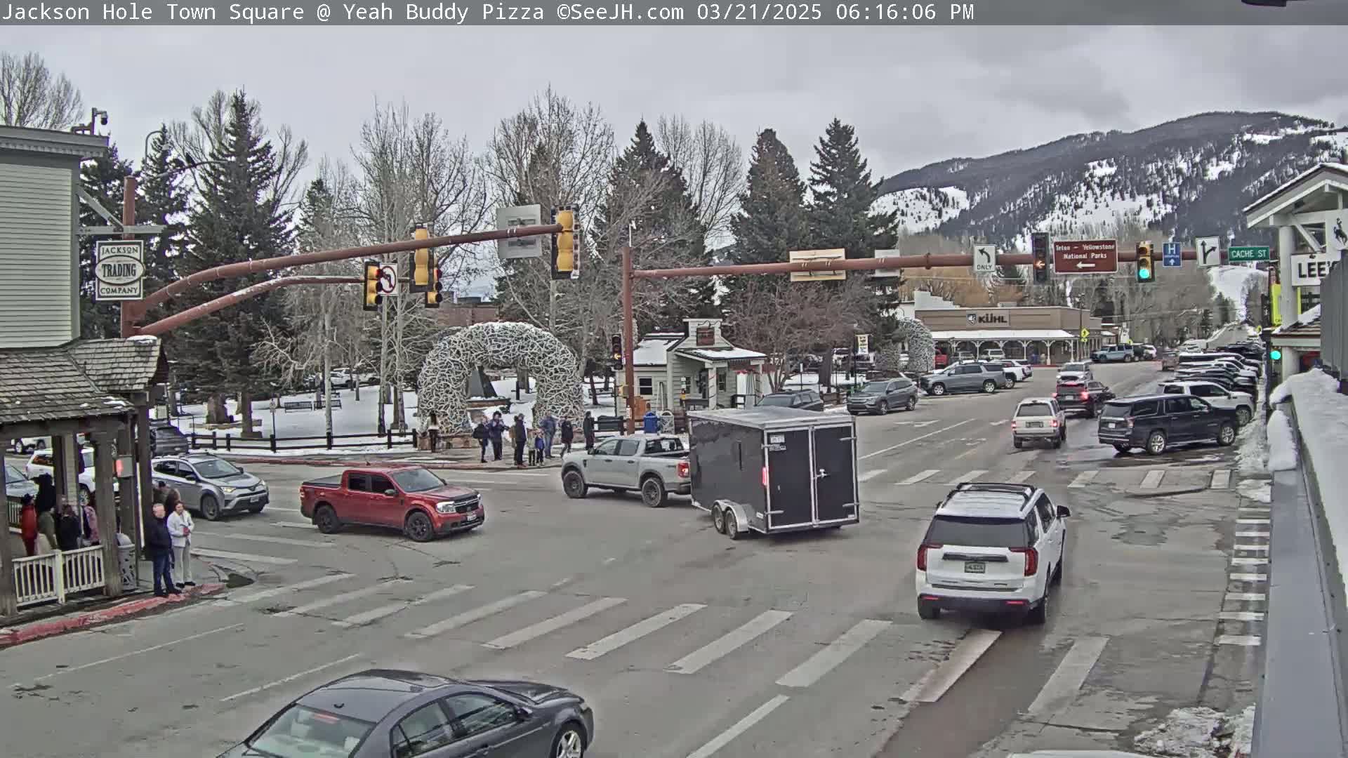 Cars are driving through a town square intersection under an overcast sky, with snow visible on the ground and mountains in the background.