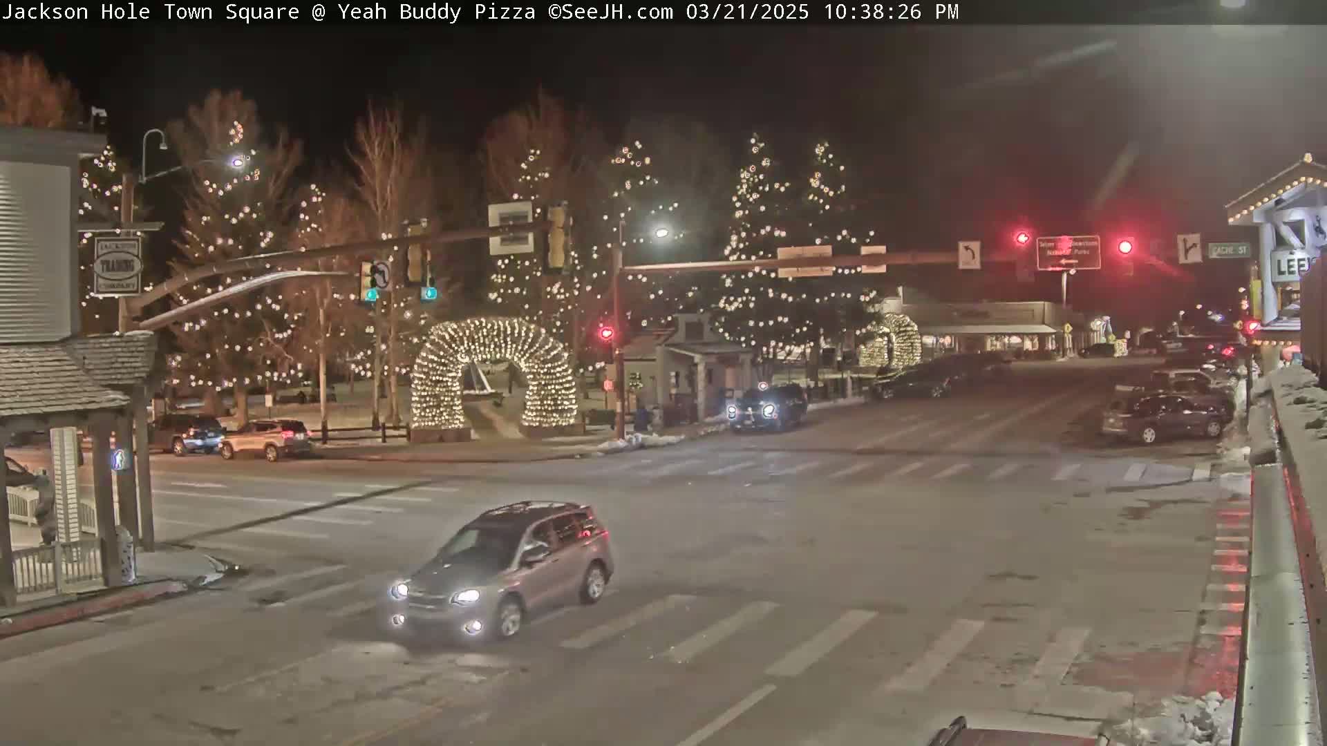 A nighttime street scene in a town square, decorated with Christmas lights on trees and an archway, shows cars at an intersection on a snowy street.