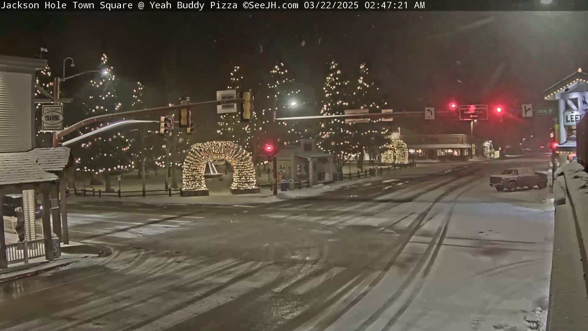 A snow-covered town square at night, illuminated by Christmas lights strung on trees and an archway, with a vehicle driving on a snow-dusted street.