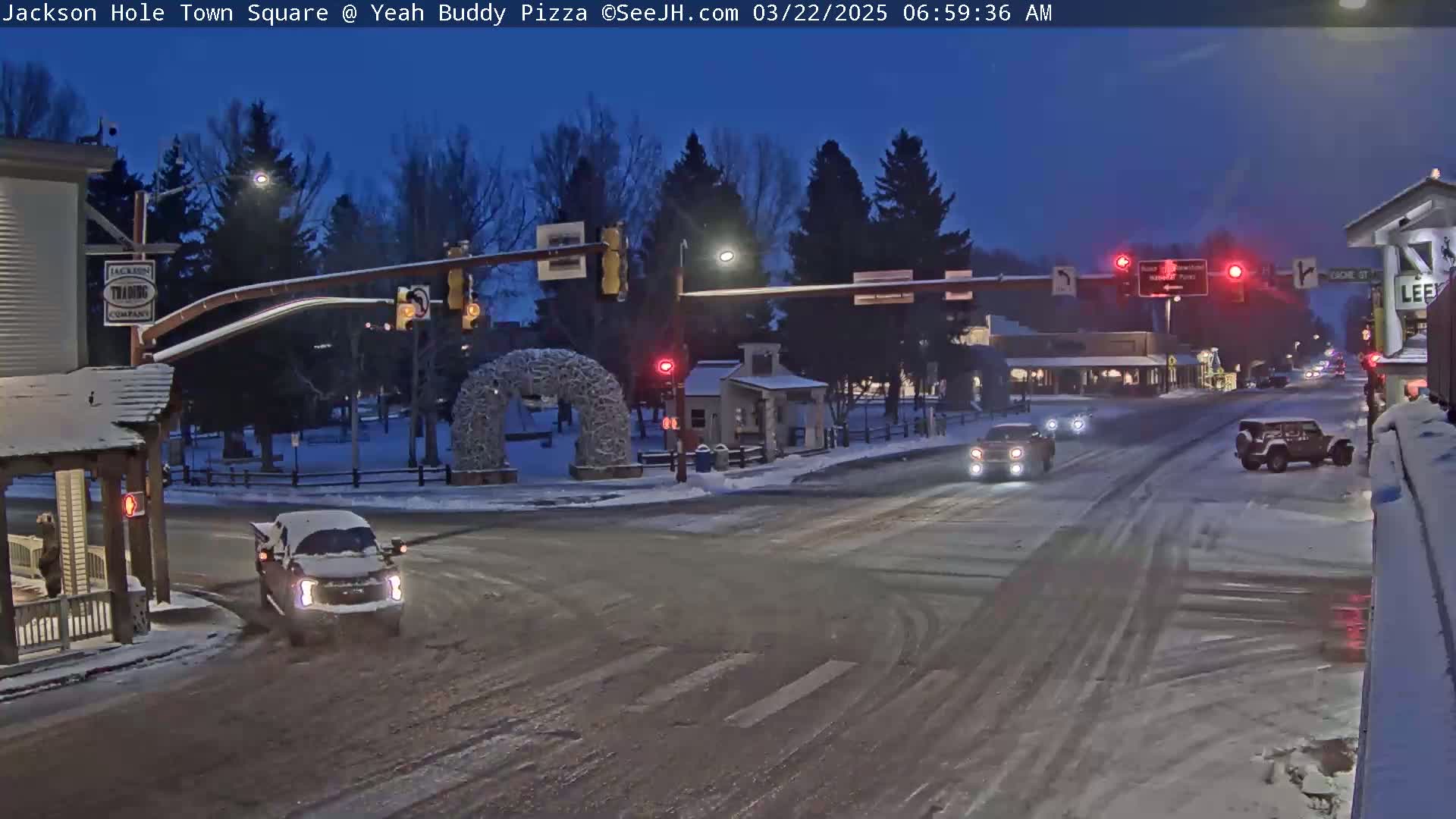 A snow-covered town square at twilight, with a few vehicles at an intersection under traffic lights.