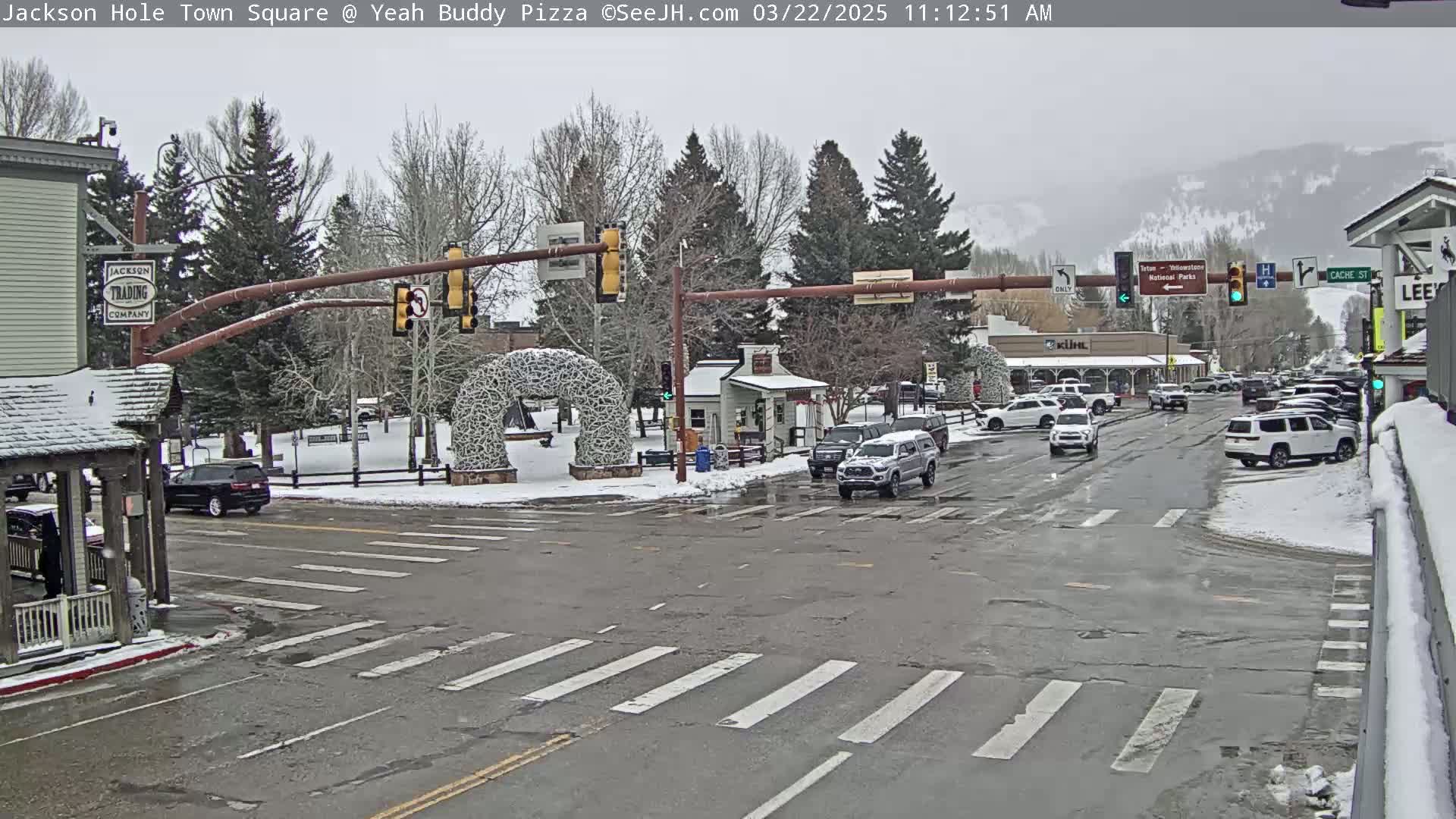 A snow-covered town square intersection with cars and traffic signals, under an overcast sky.