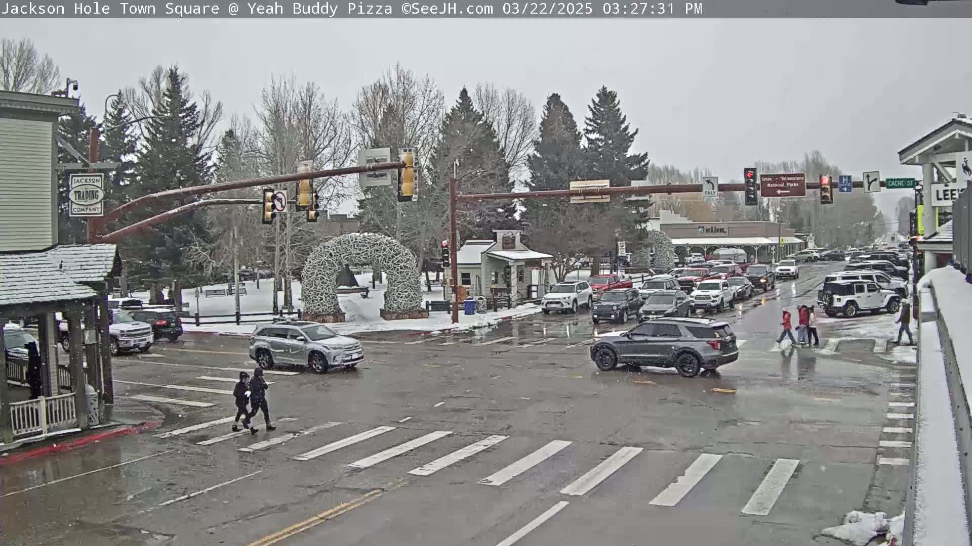 A snowy town square intersection shows several vehicles stopped at traffic signals, with pedestrians crossing the street in light snowfall.