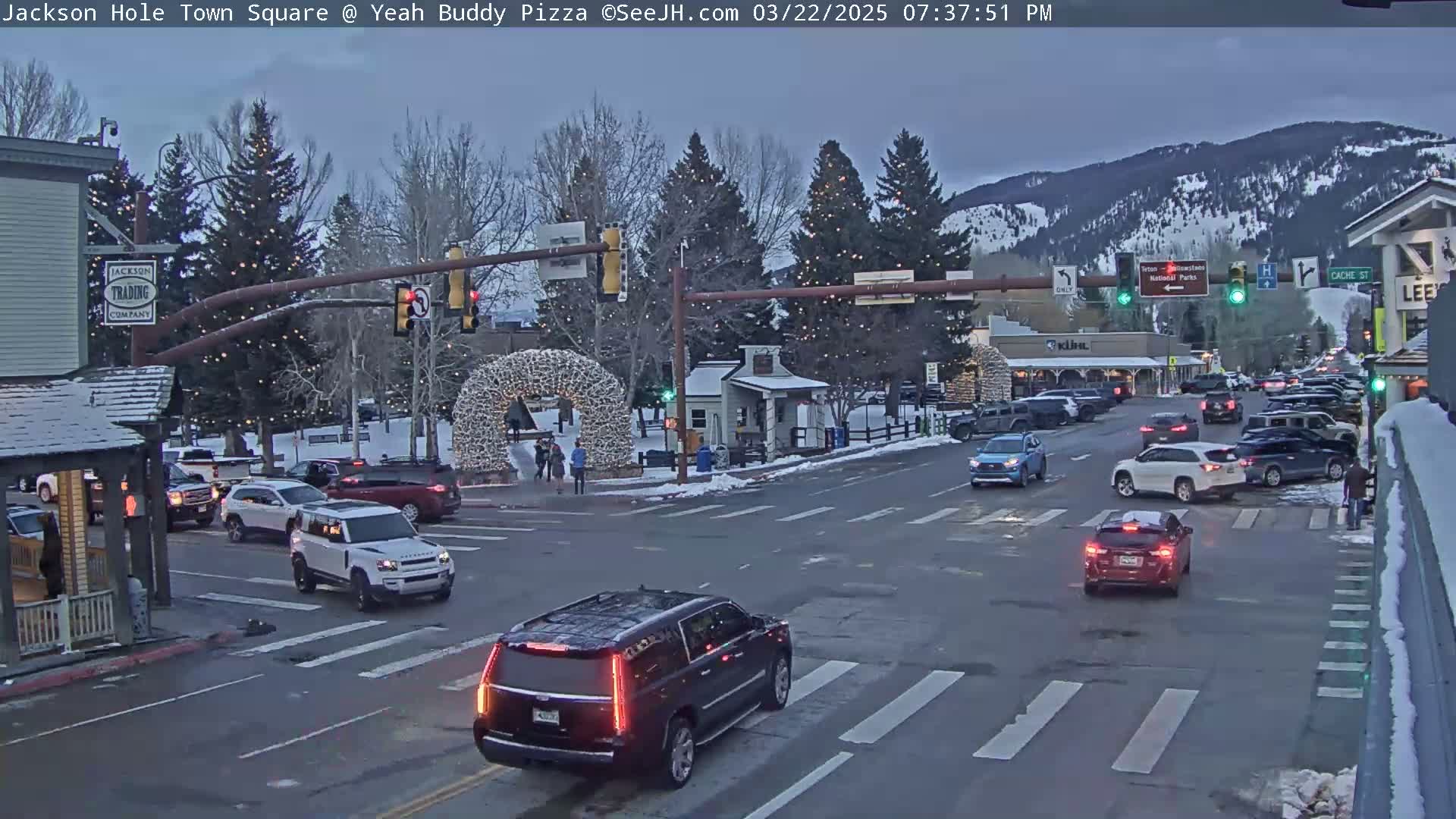 A town square at dusk with several vehicles at an intersection, under partly cloudy skies, with snow on the ground in some areas and Christmas lights on trees.