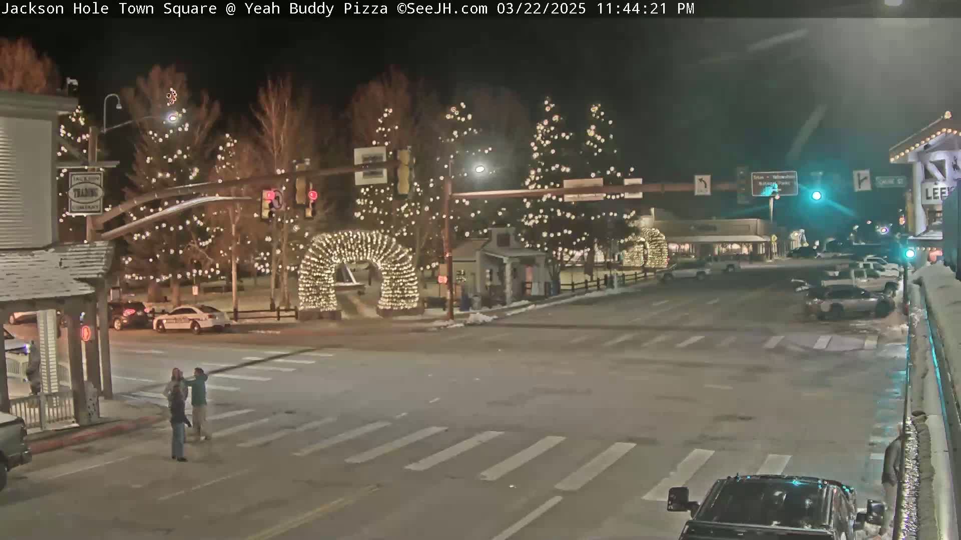 A nighttime street scene shows a town square decorated with Christmas lights, with a few cars and pedestrians present on a snowy street.