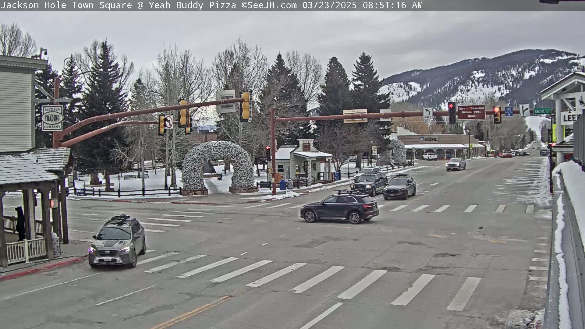 A town square intersection with several vehicles, snow-covered ground, and bare trees under an overcast sky.