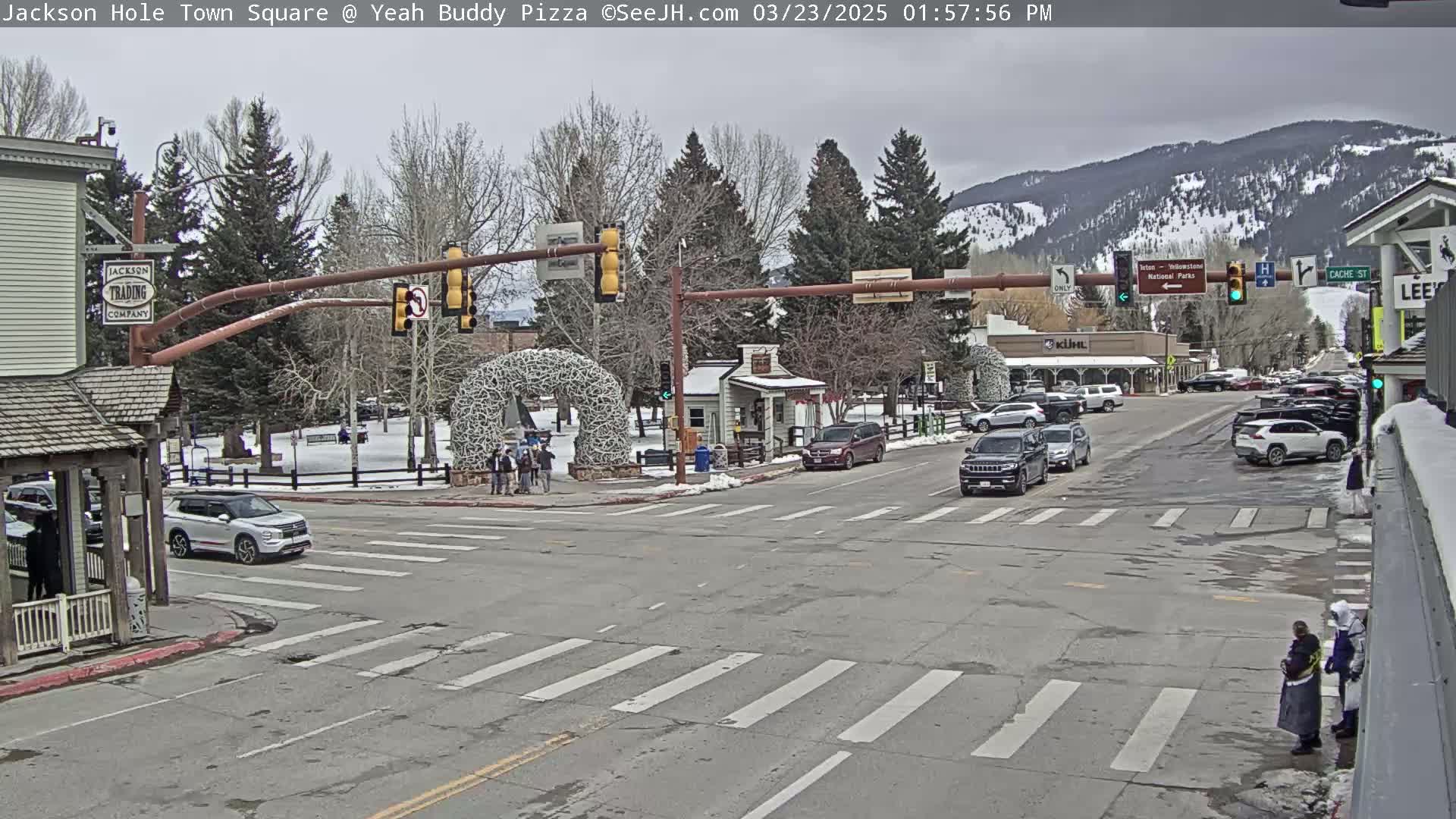 A town square intersection with cars and pedestrians, under an overcast sky with snow on the ground and mountains in the background.
