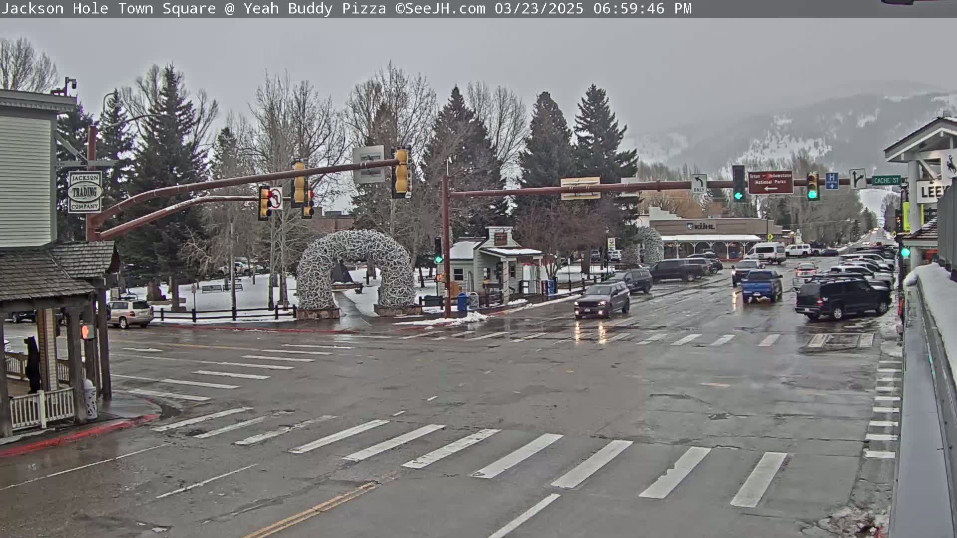 A wet town square intersection with traffic, snow on the ground, and bare, leafless trees under a gray sky.