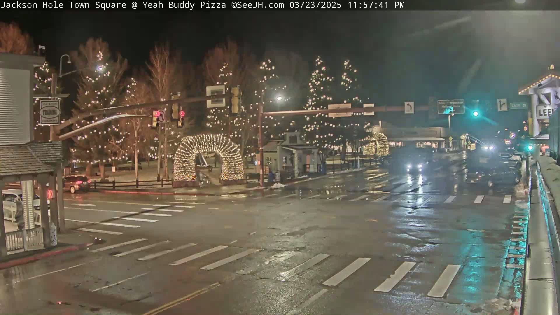 A town square at night, decorated with Christmas lights, is wet from recent rain or snowmelt, with several cars driving through the intersection.