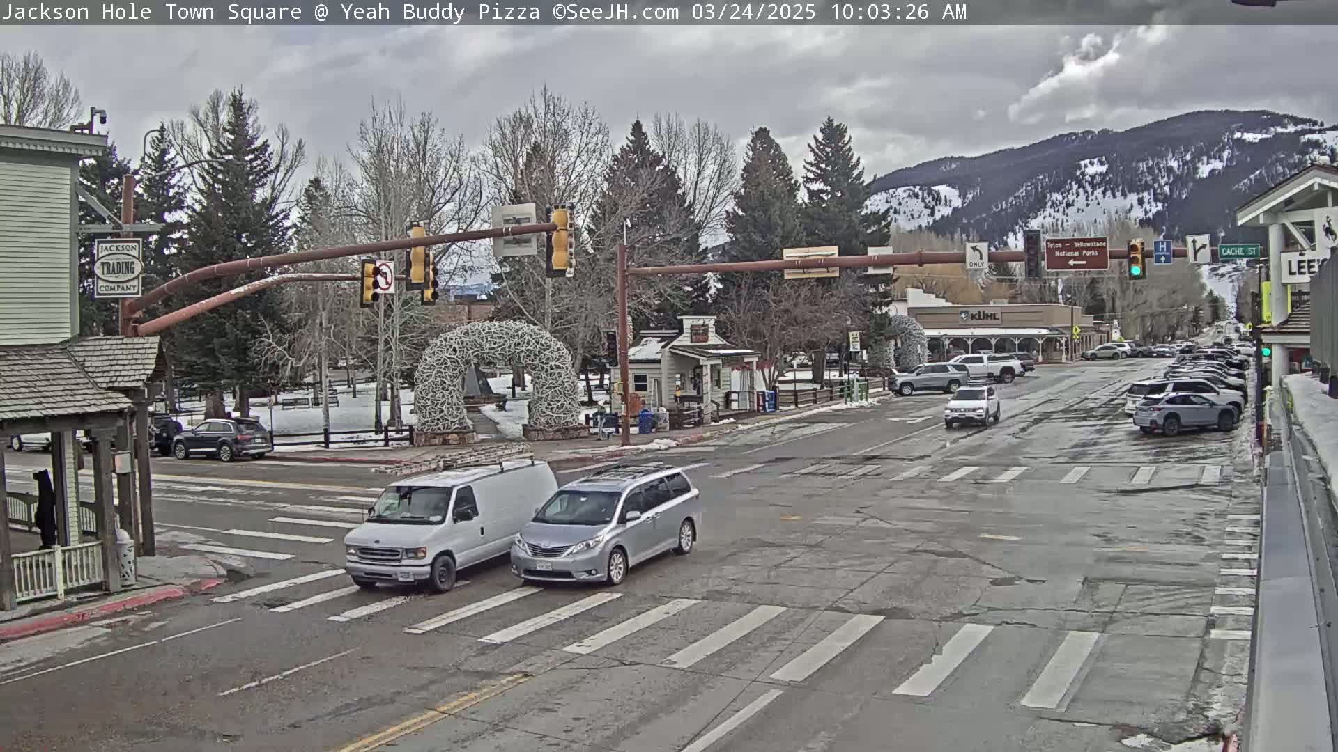 A mostly cloudy day in a town square shows several vehicles at an intersection with snow on the ground and mountains in the background.