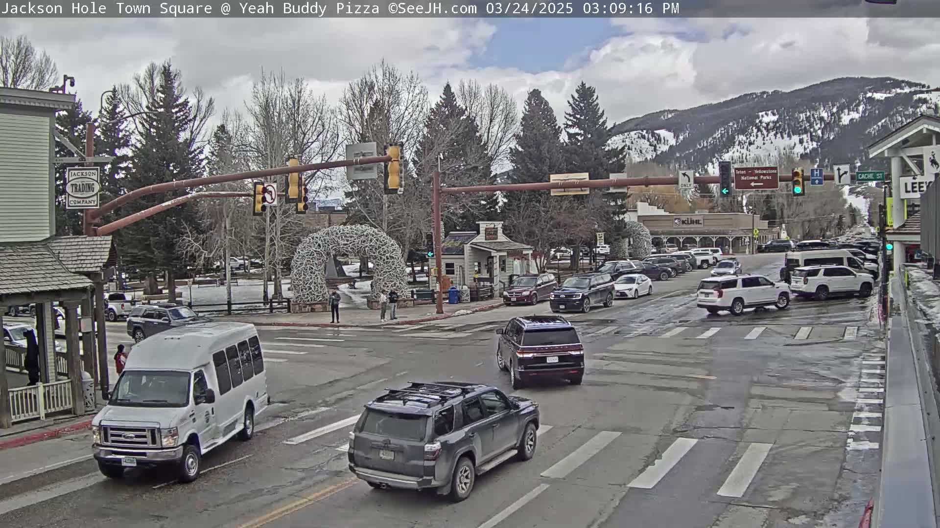 A town square with several cars at an intersection under a partly cloudy sky, with mountains in the background.