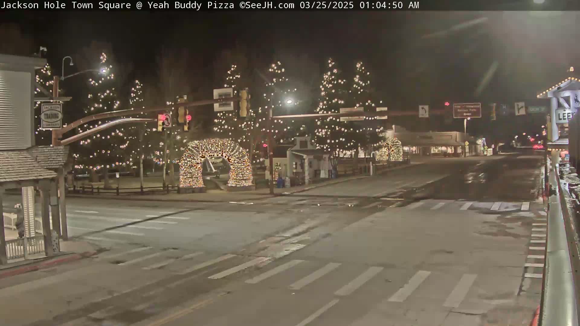 A nighttime view of a town square with Christmas lights decorating trees and arches, and wet roads.