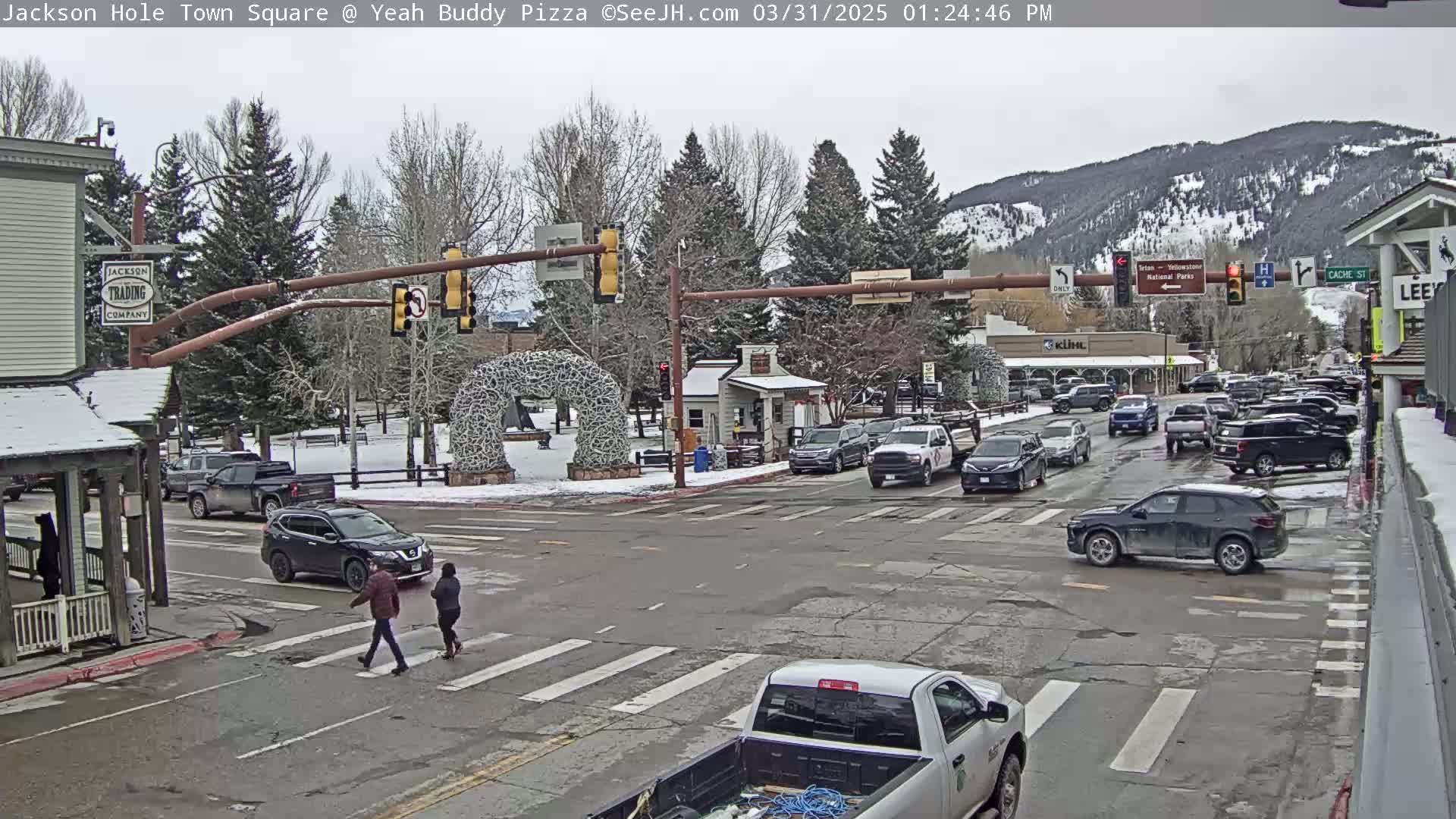A town square intersection with several cars and pedestrians, under an overcast sky, with snow on the ground and mountains in the background.